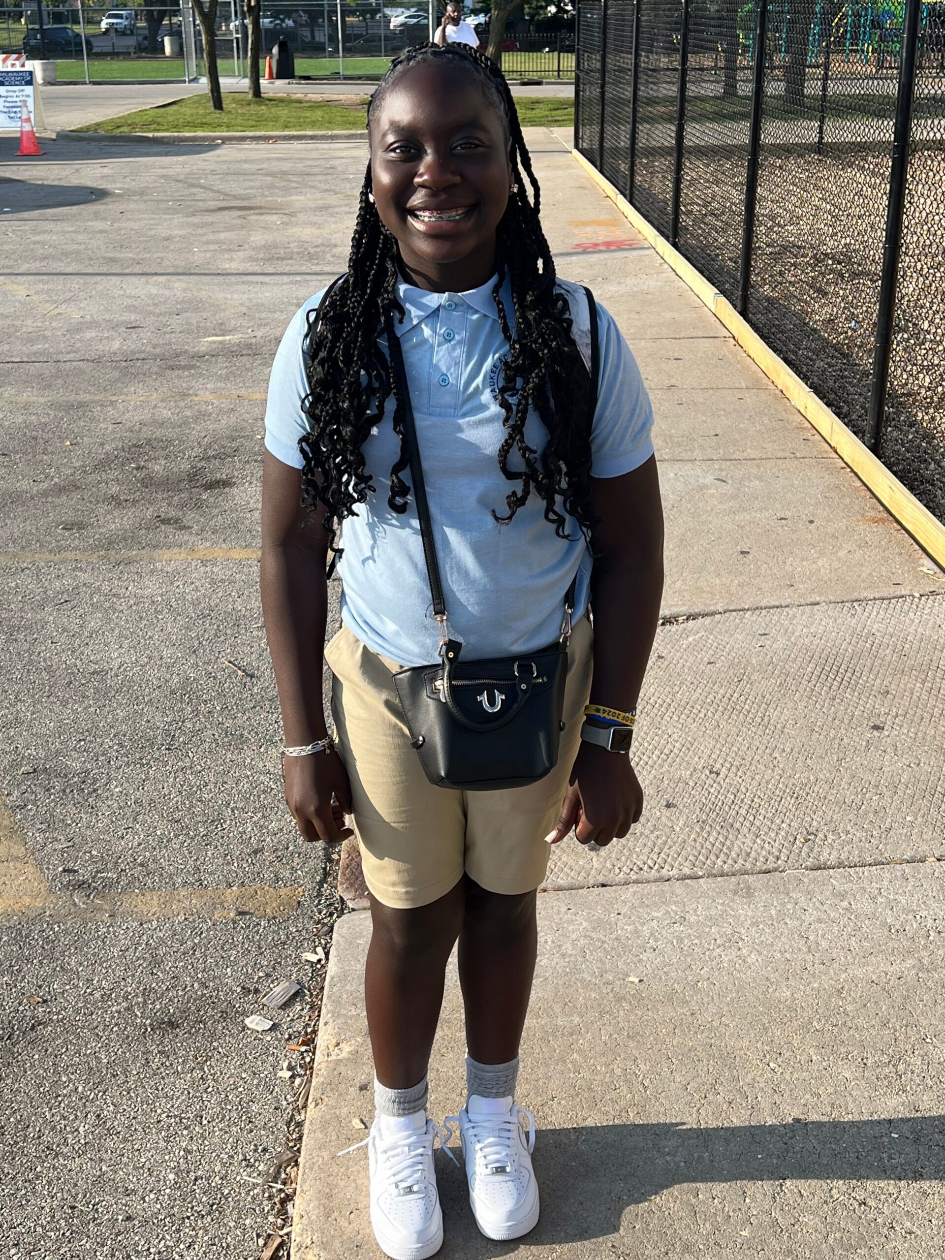 Young girl smiling at camera, wearing a light blue polo, khaki shorts, white sneakers, and a small black crossbody bag, outdoors near a fence.