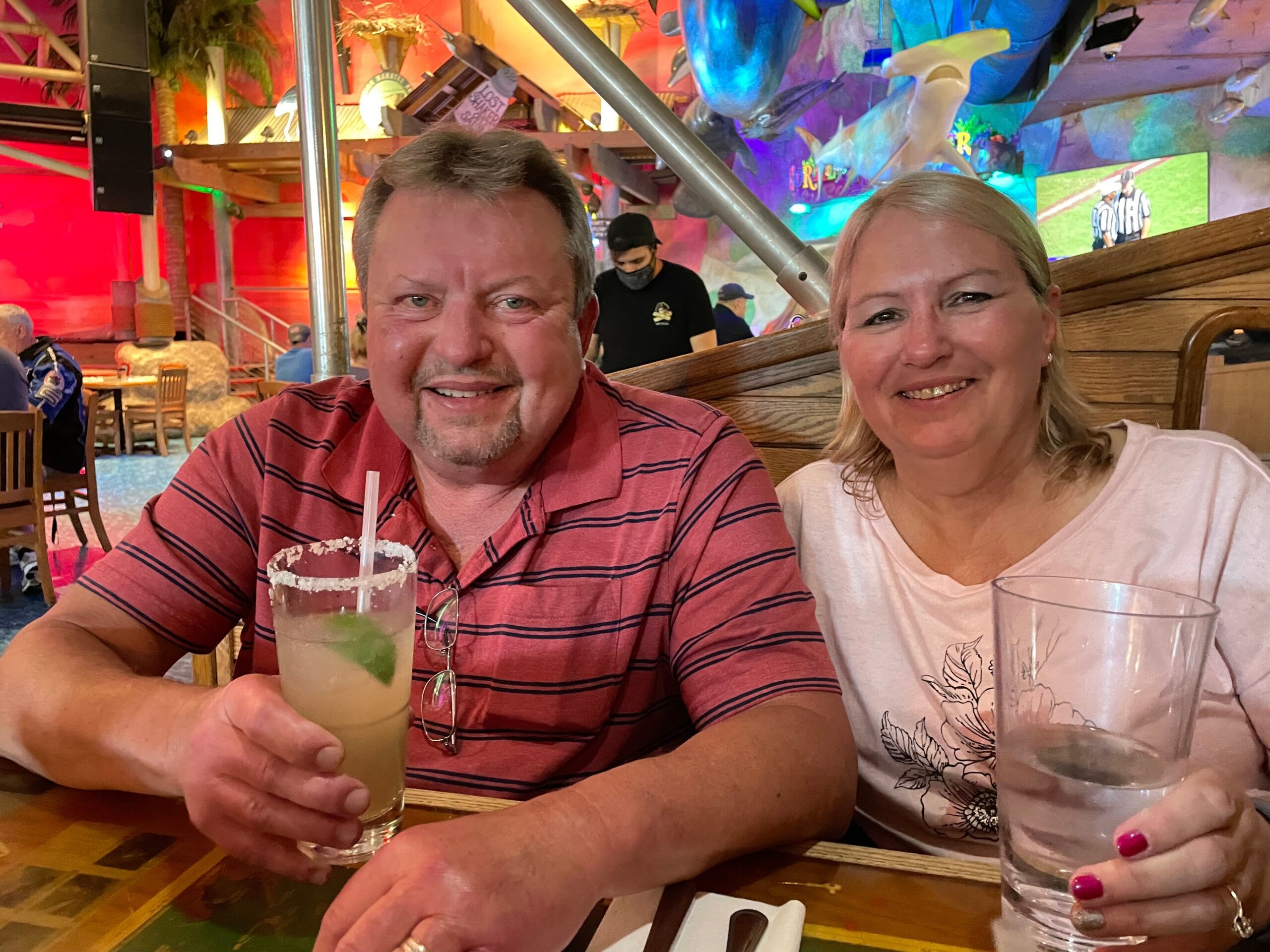 Smiling man and woman sit at a table with drinks in a vibrant, mural-filled restaurant backdrop.