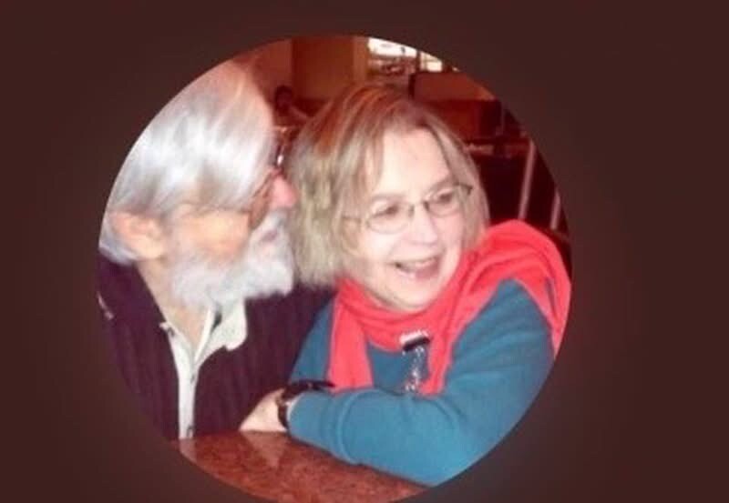 Smiling elderly couple sitting at a table, leaning toward each other in a cozy setting.
