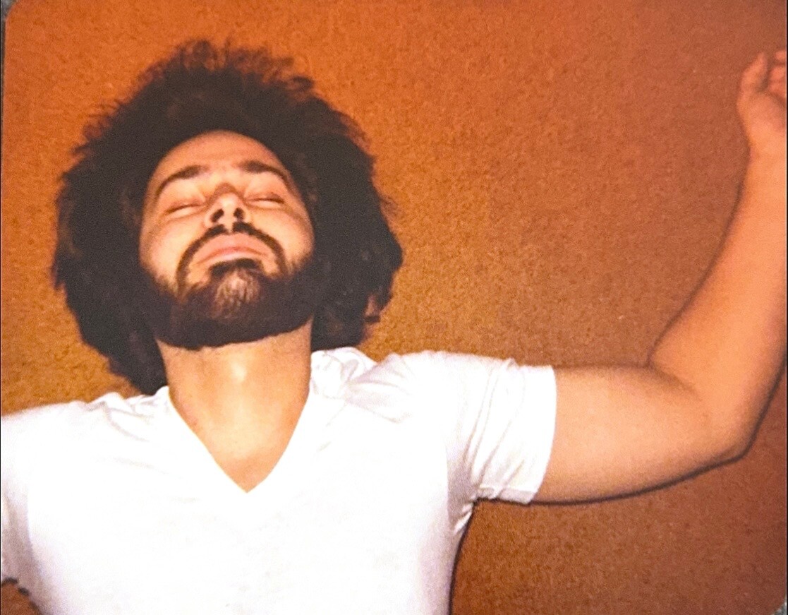 Bearded man with eyes closed and arms raised, wearing a white shirt, standing against an orange wall.