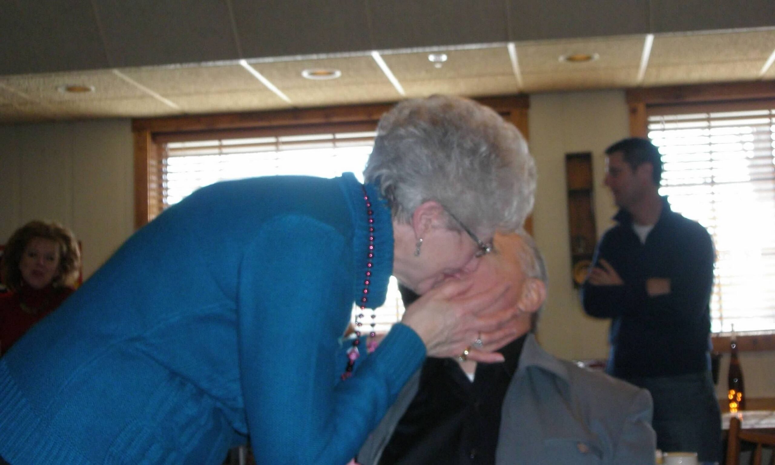 Woman in a blue sweater leans in to kiss an elderly person on the cheek at a family gathering, with a man standing in the background indoors.