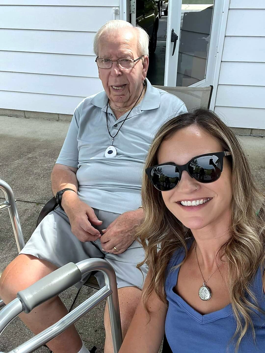 Smiling woman in a blue top taking a selfie with an elderly man in a light blue polo seated outside a house; a walker is in the foreground.