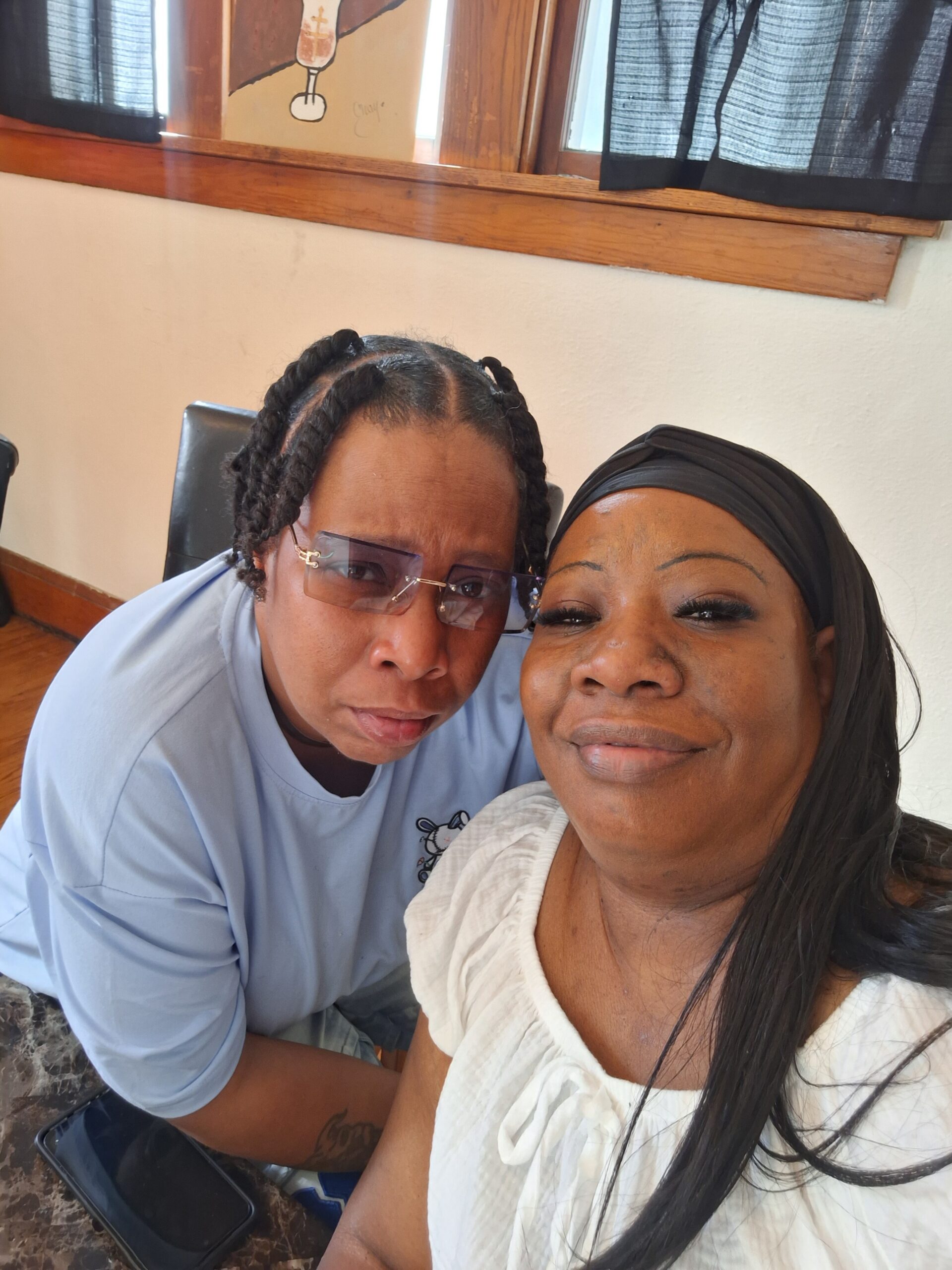 Close-up selfie of two smiling women indoors: one with braids and tinted glasses, the other wearing a white blouse and black headscarf.