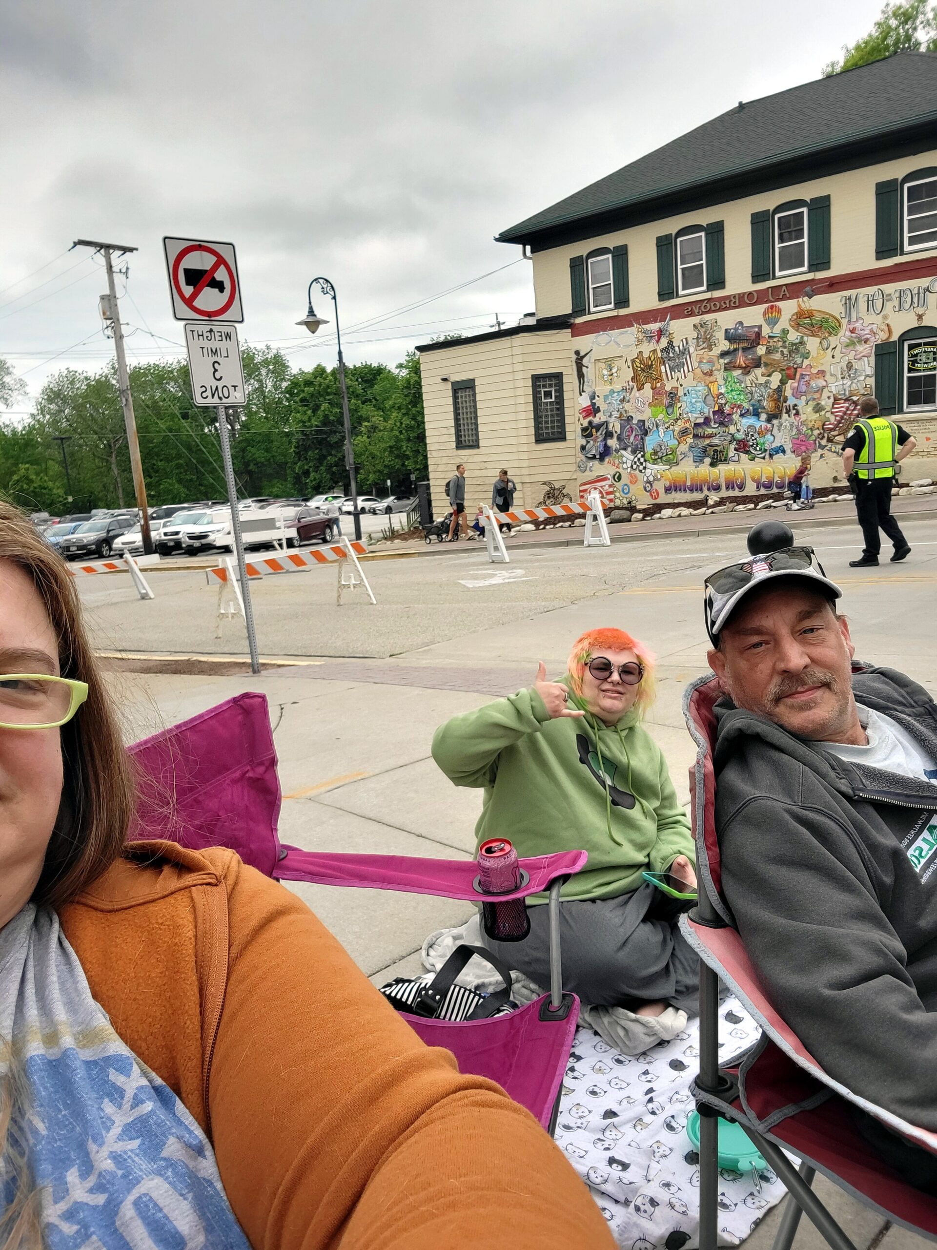 A group of people sitting and taking a selfie on a city street, with a colorful mural on a building behind them and parked cars in the background.
