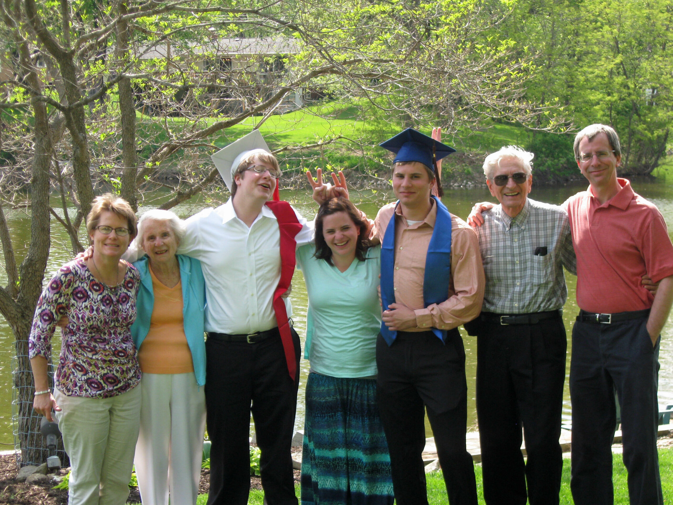 Six people posing together outdoors by a lake on a sunny day; two graduates wear caps and sashes among friends and family.