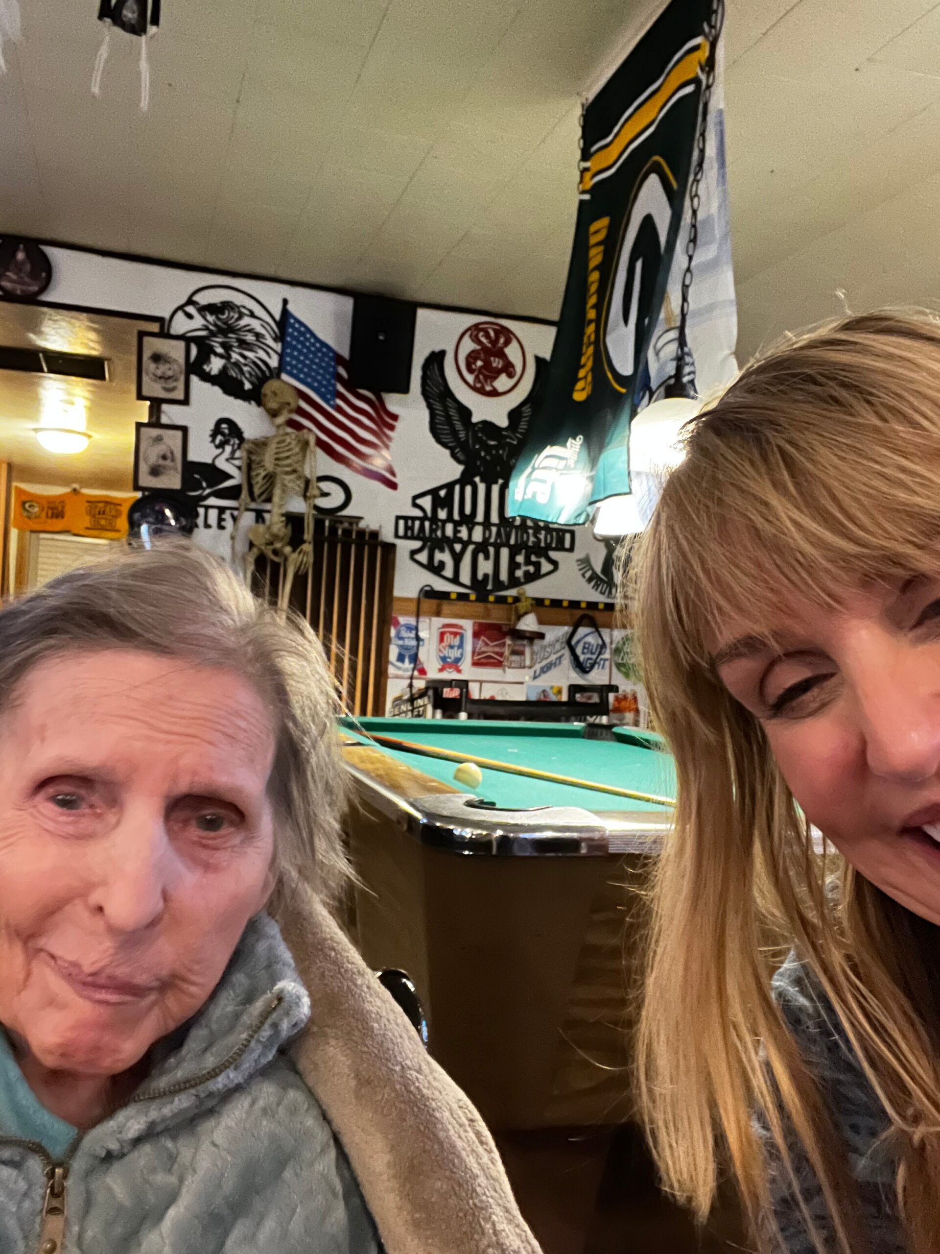 Two people taking a casual selfie in a biker-themed room with Harley‑Davidson wall art, an American flag, and a hanging banner above a pool table behind them.