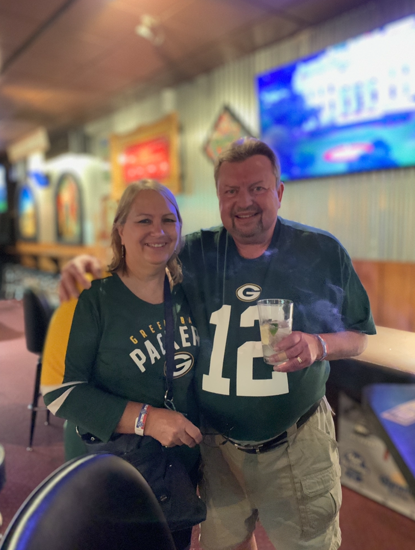 Smiling couple posing together in a bar, both wearing Green Bay Packers attire; the man holds a drink.