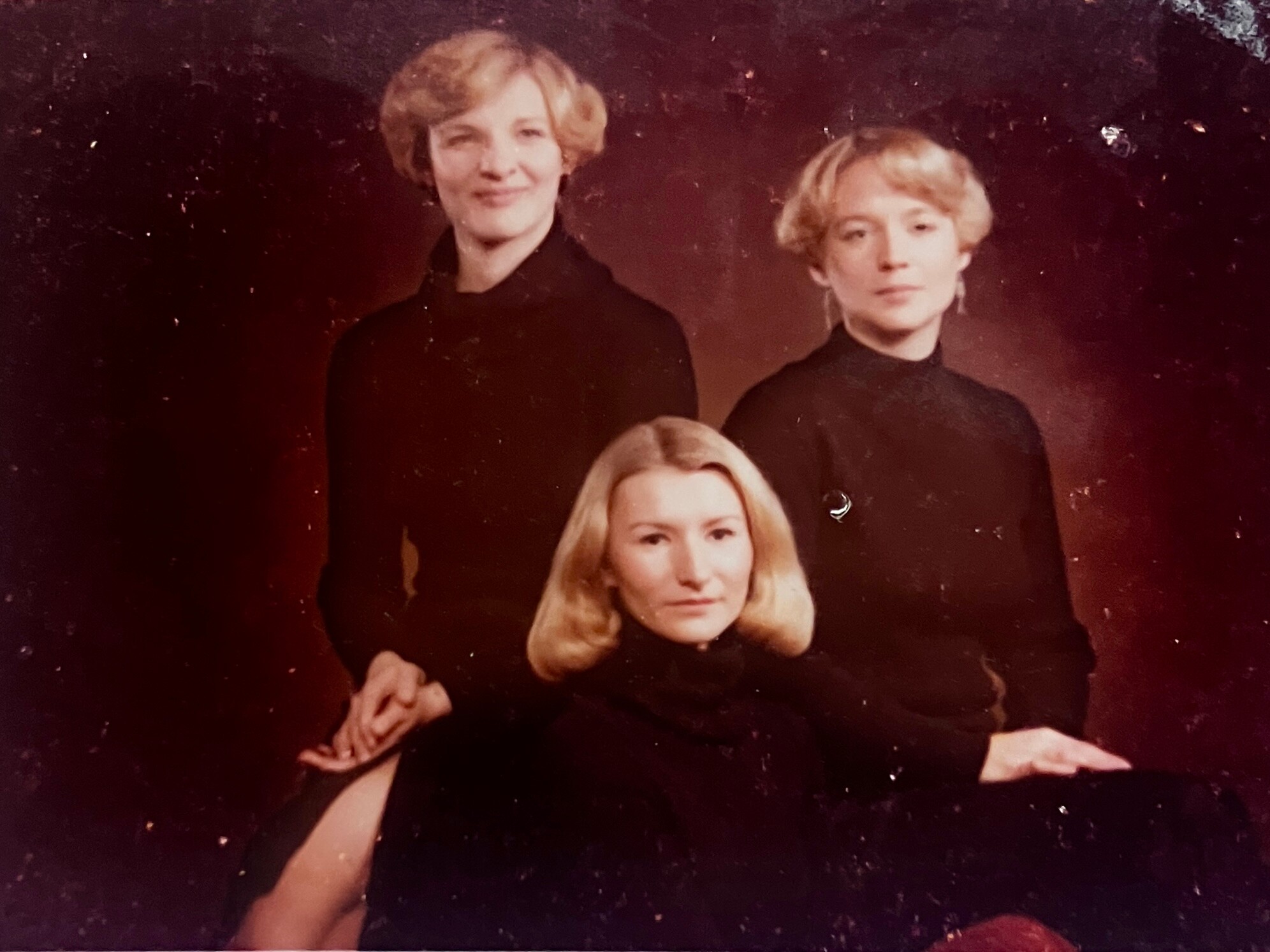 Three women in black turtlenecks posing for a studio portrait, with two behind and one in front.