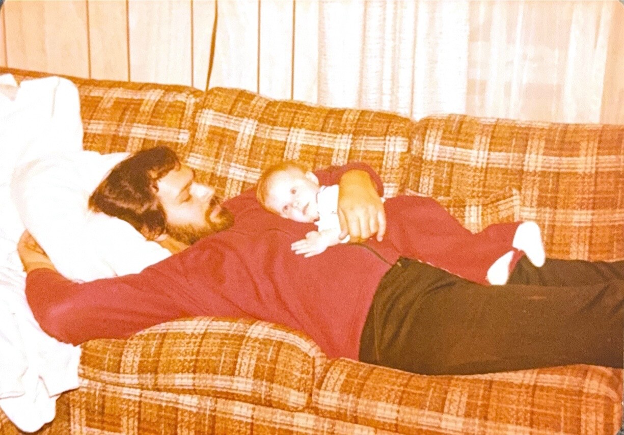 Man with a beard lying on a plaid orange couch, cradling a baby on his chest.