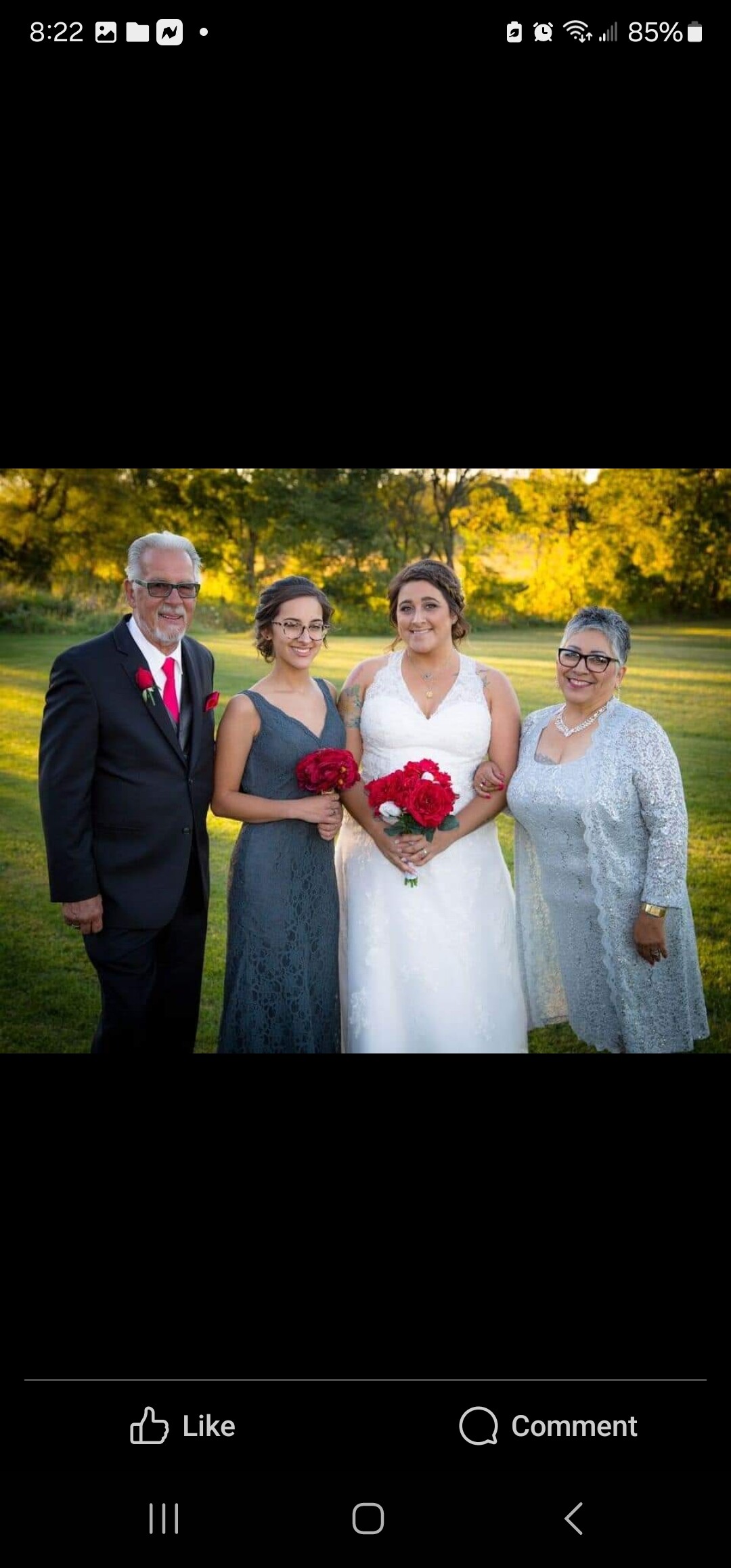 Wedding party outdoors: bride in white holding red roses, flanked by two bridesmaids in gray dresses and an older man in a dark suit with a red tie on a sunlit lawn.