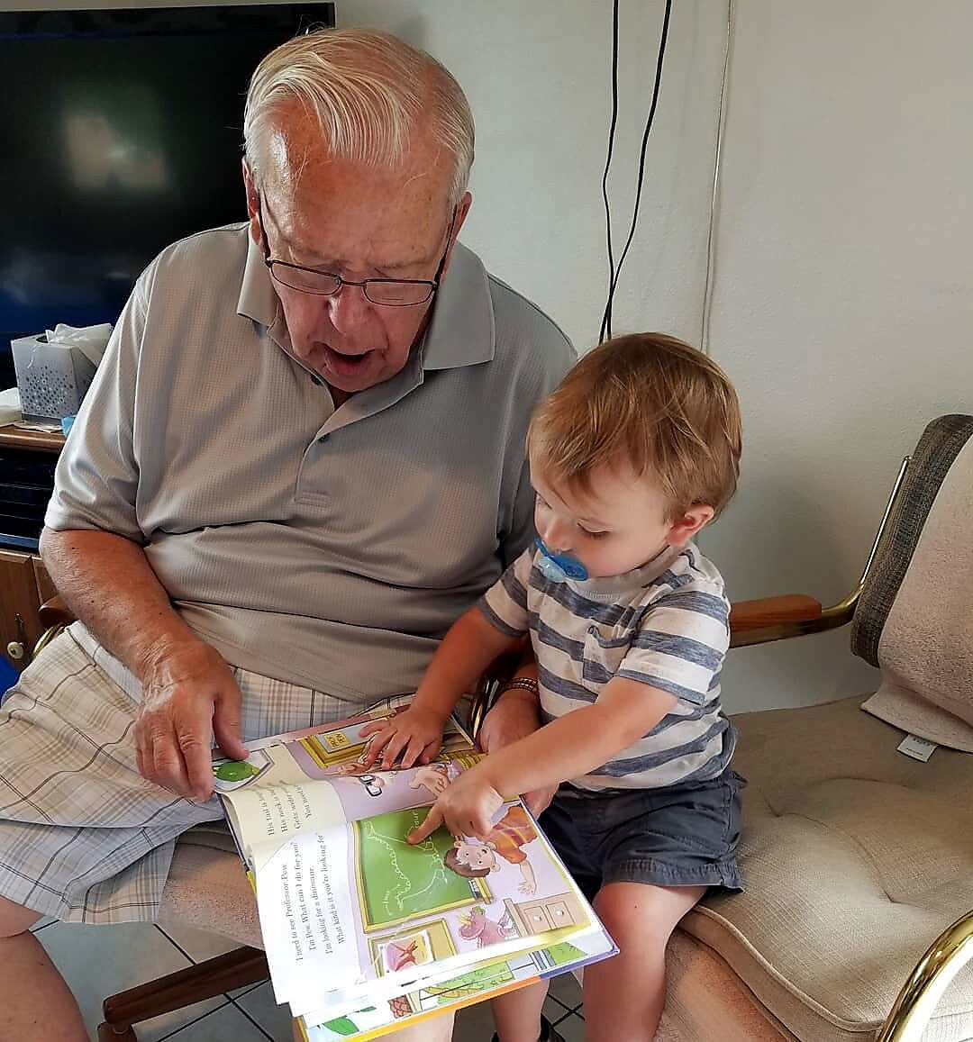 Grandfather reading a colorful children's book to a toddler with a blue pacifier.