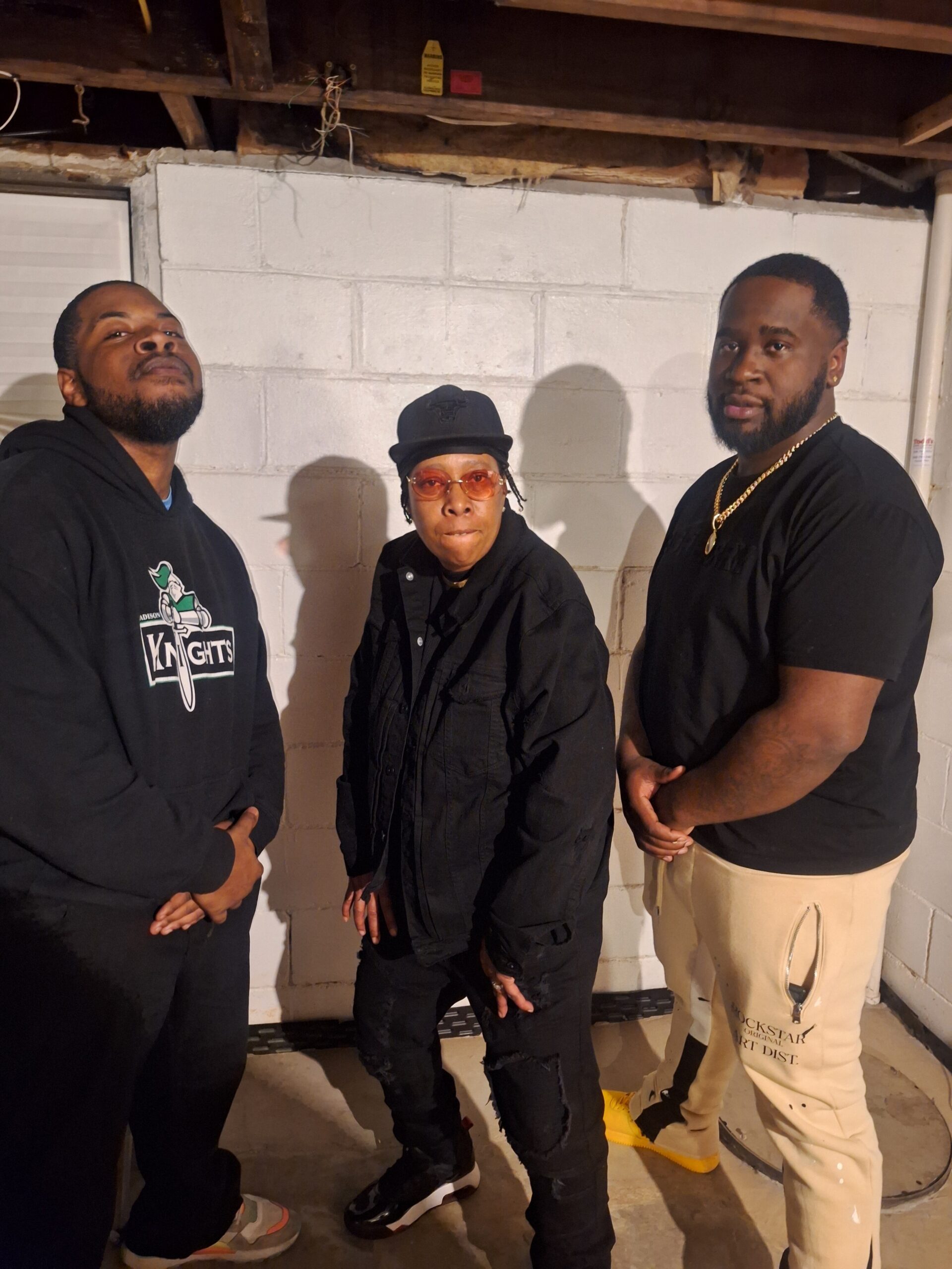 Three people pose in a basement against a white cinder block wall: left man in a black hoodie with a Knights graphic, middle woman in a black jacket and hat with orange glasses, right man in a black shirt with a gold chain.