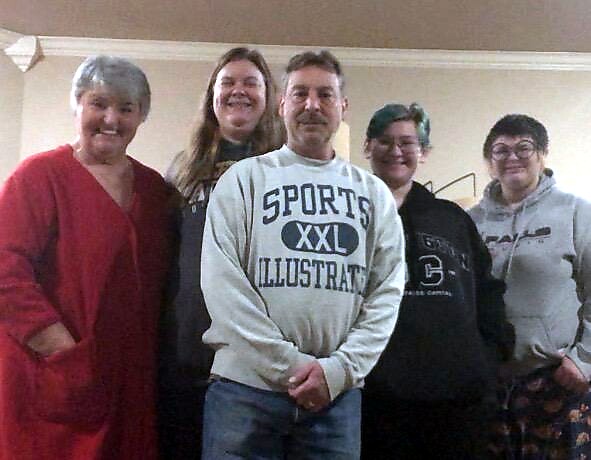 Six adults indoors posing for a photo; the man in the center wears a light gray sweatshirt reading 'SPORTS ILLUSTRATED XXL' and others smile around him.