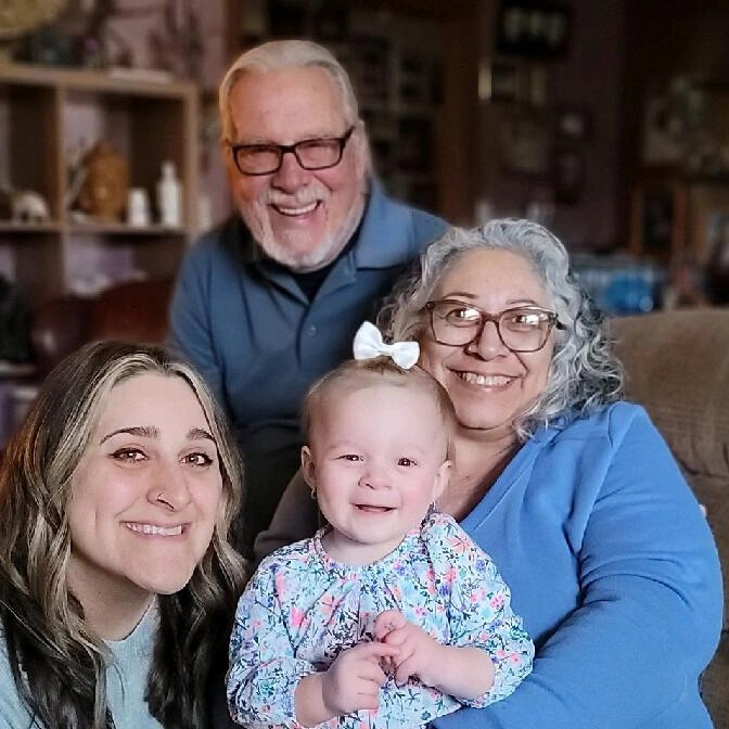 Four generations smiling together on a couch: a baby girl in front, her mother, grandmother, and grandfather behind them in a cozy living room.