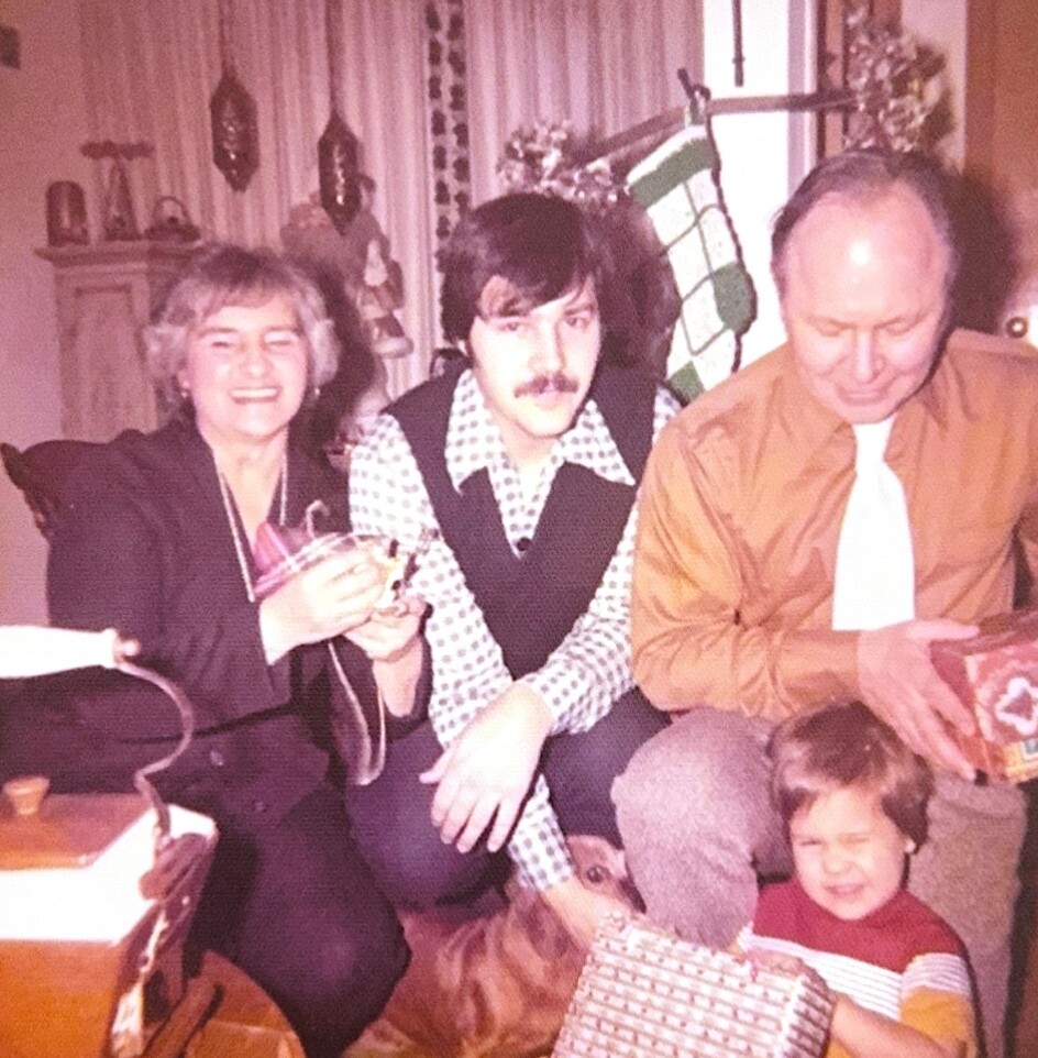 Smiling woman and two men sit together during a Christmas gathering, with a child opening presents on the floor nearby.
