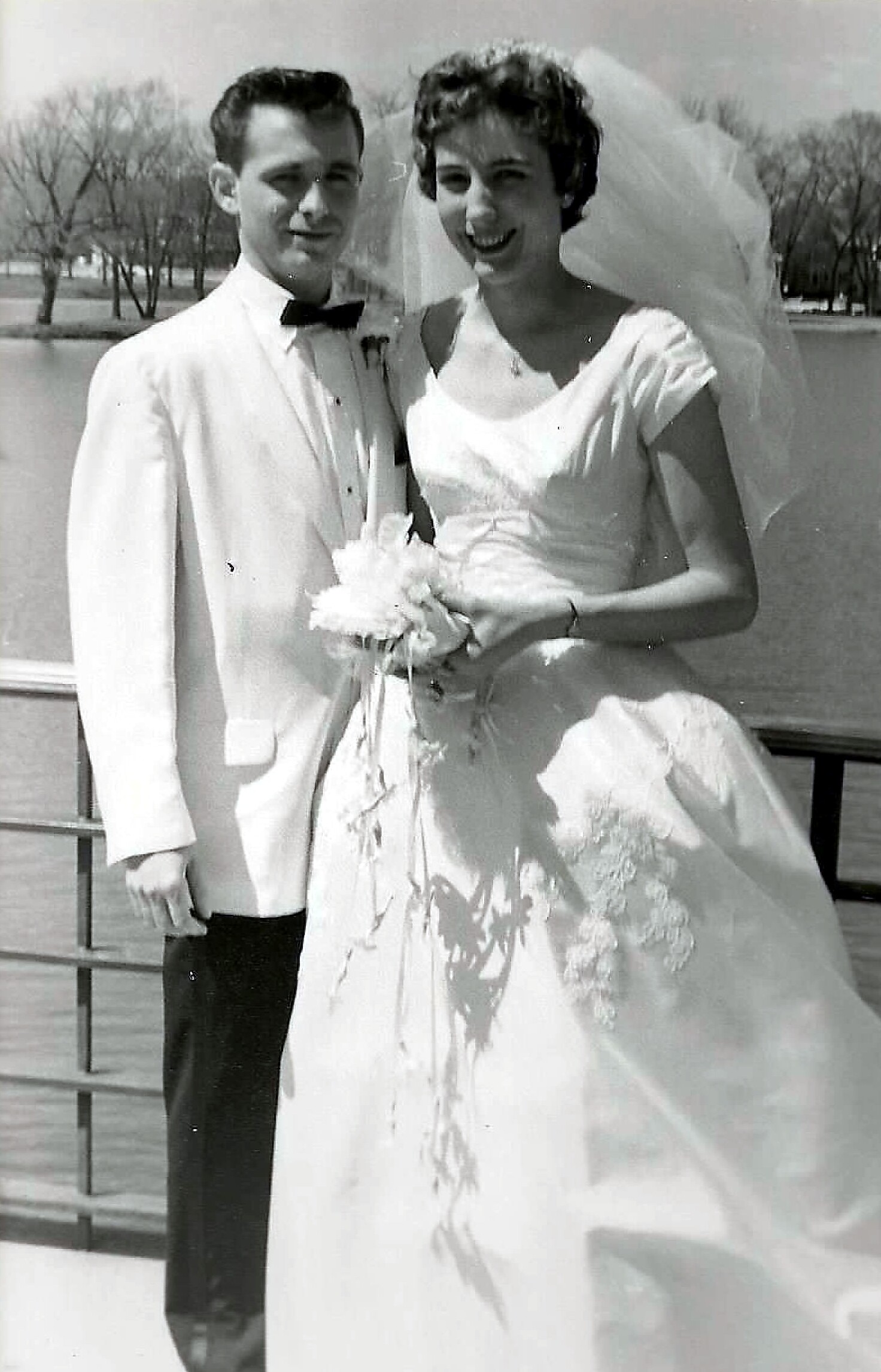 Bride in a white wedding gown with veil holding a bouquet, standing beside a groom in a light tuxedo by a railing near a lake, both smiling.