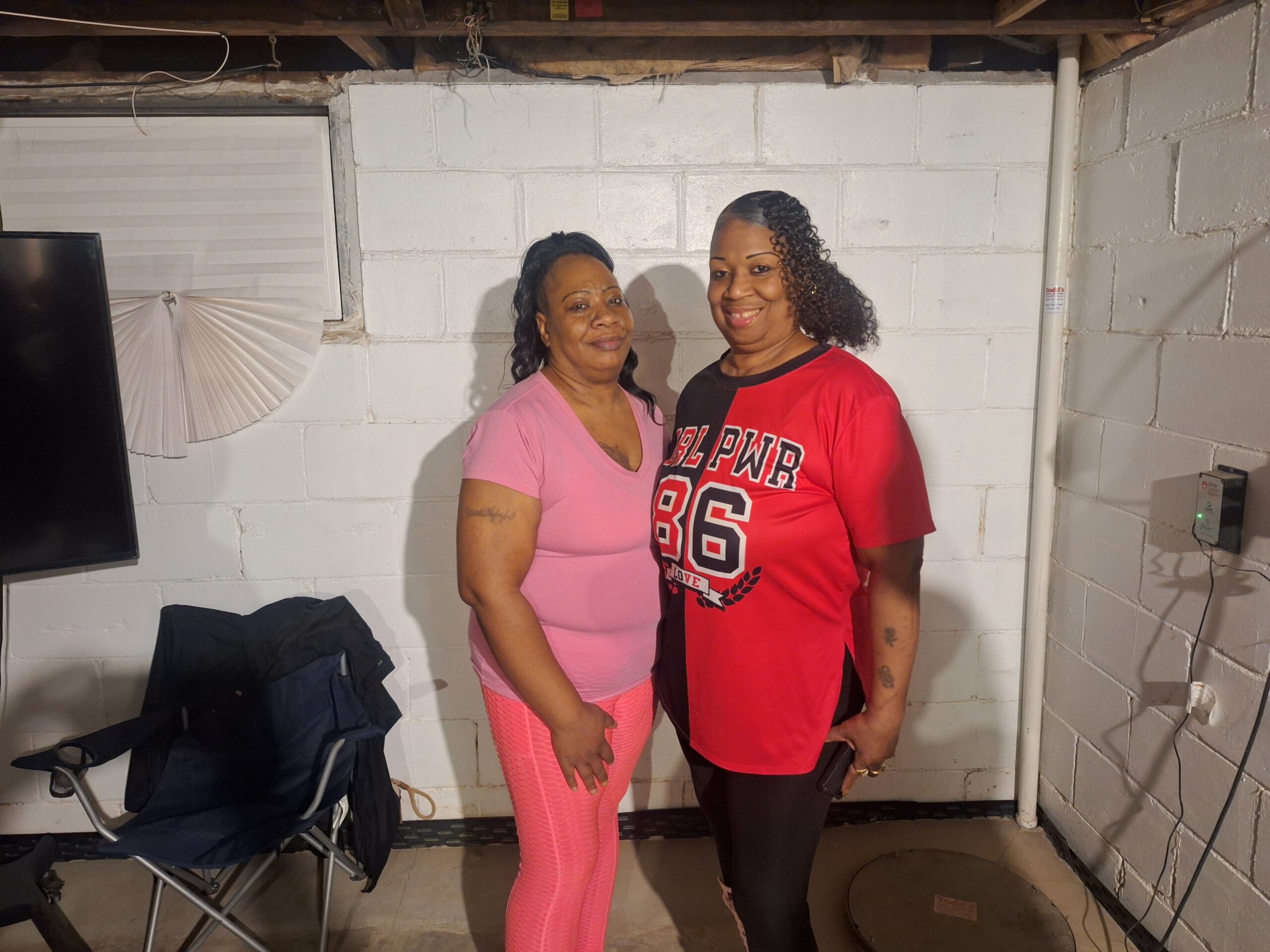 Two women stand side by side in a basement, smiling at the camera; one in pink athletic wear and the other in a red and black sports jersey.
