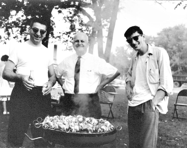 Three men stand around a charcoal grill outdoors, smiling as they cook skewers. (Black-and-white photo)