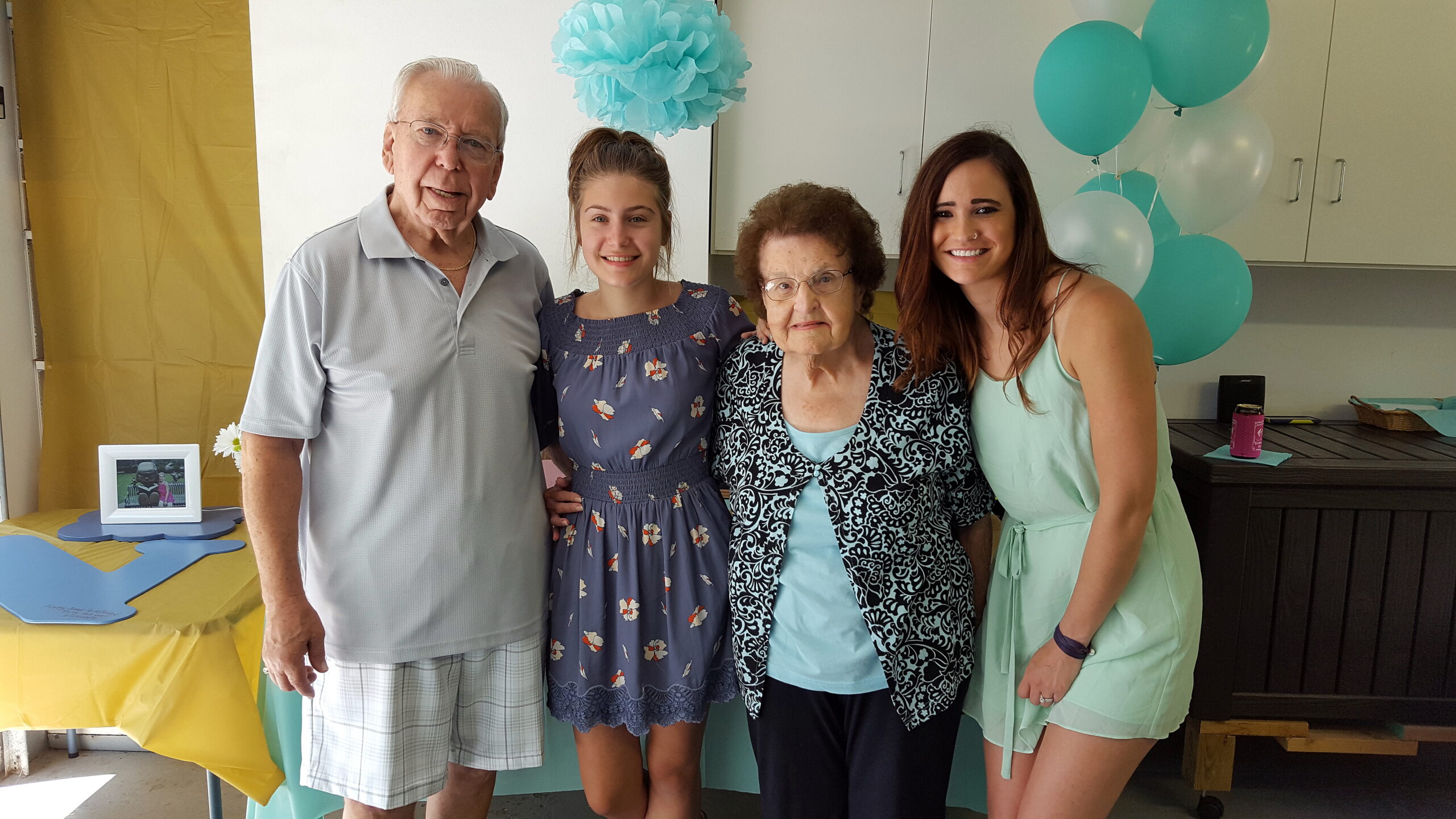 Four people posing together at a celebration with teal balloons and a yellow tablecloth in the background.