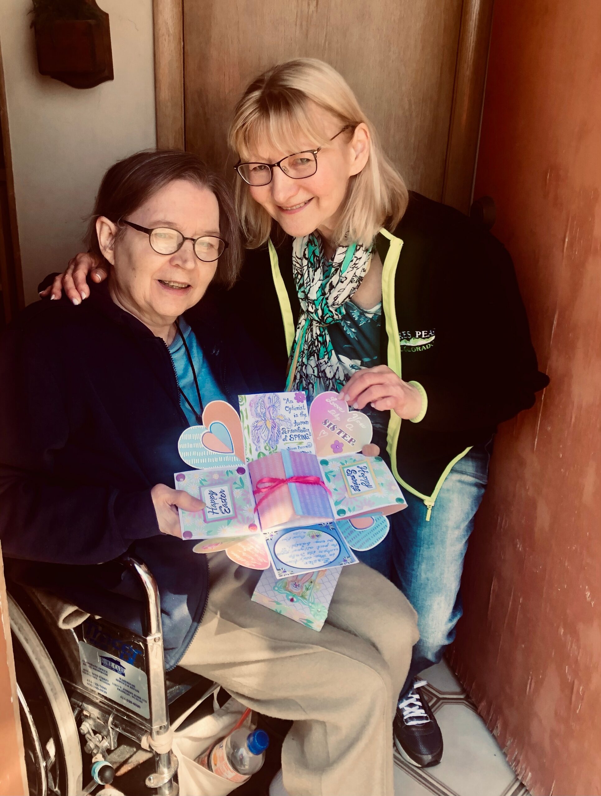 Two smiling women sit closely in a small elevator or booth, holding colorful cards and gifts displayed together.