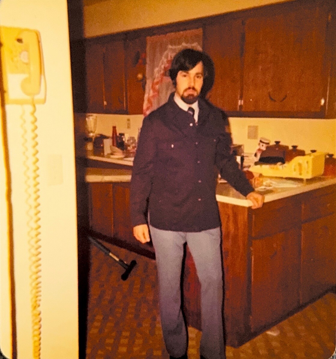 Man wearing a dark buttoned jacket and light trousers stands in a retro-style kitchen with wooden cabinets and cluttered counters.