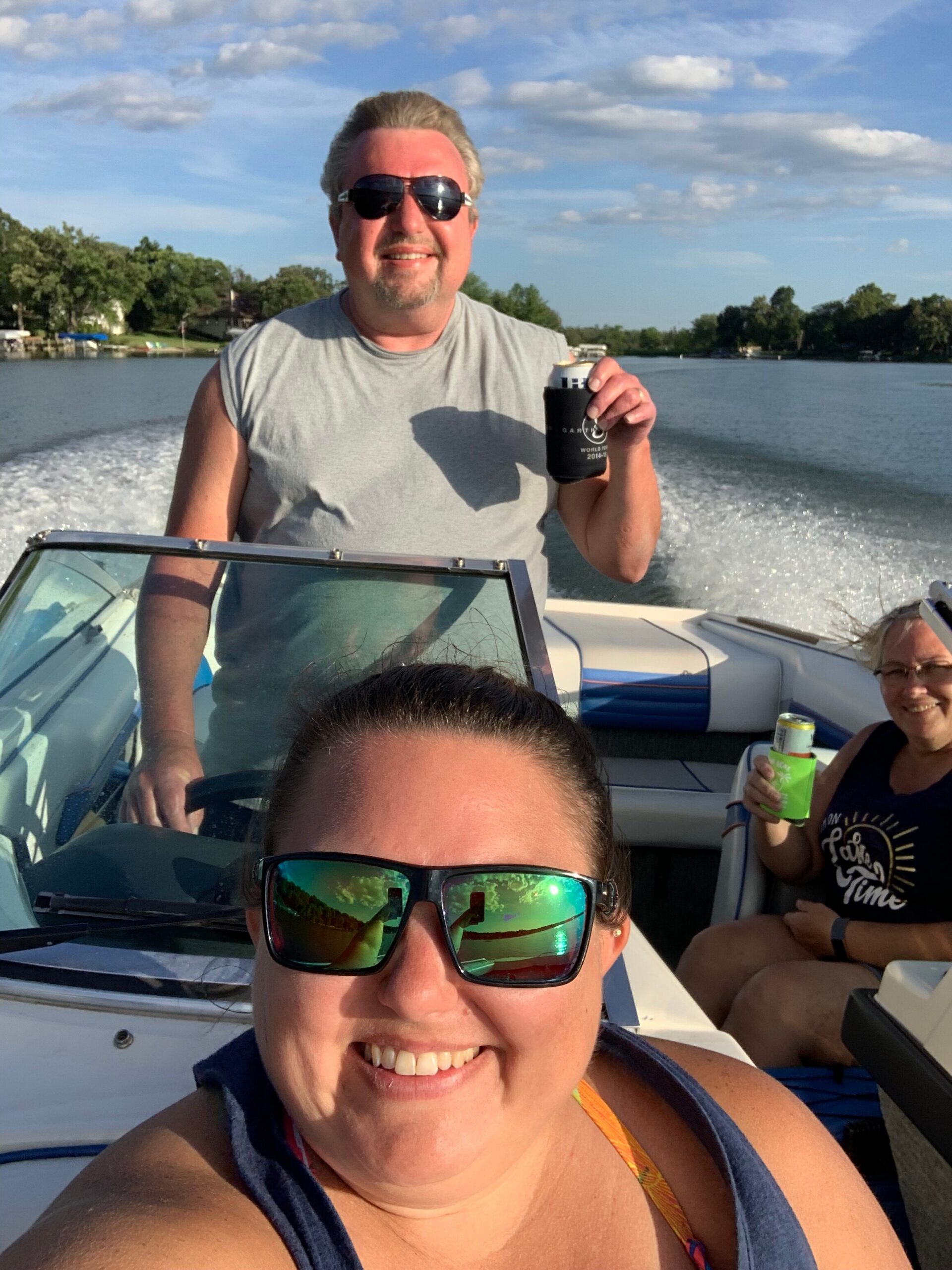 Person in the foreground takes a selfie on a boat, smiling, with two others and water in the background; man behind holds a can and wears sunglasses.