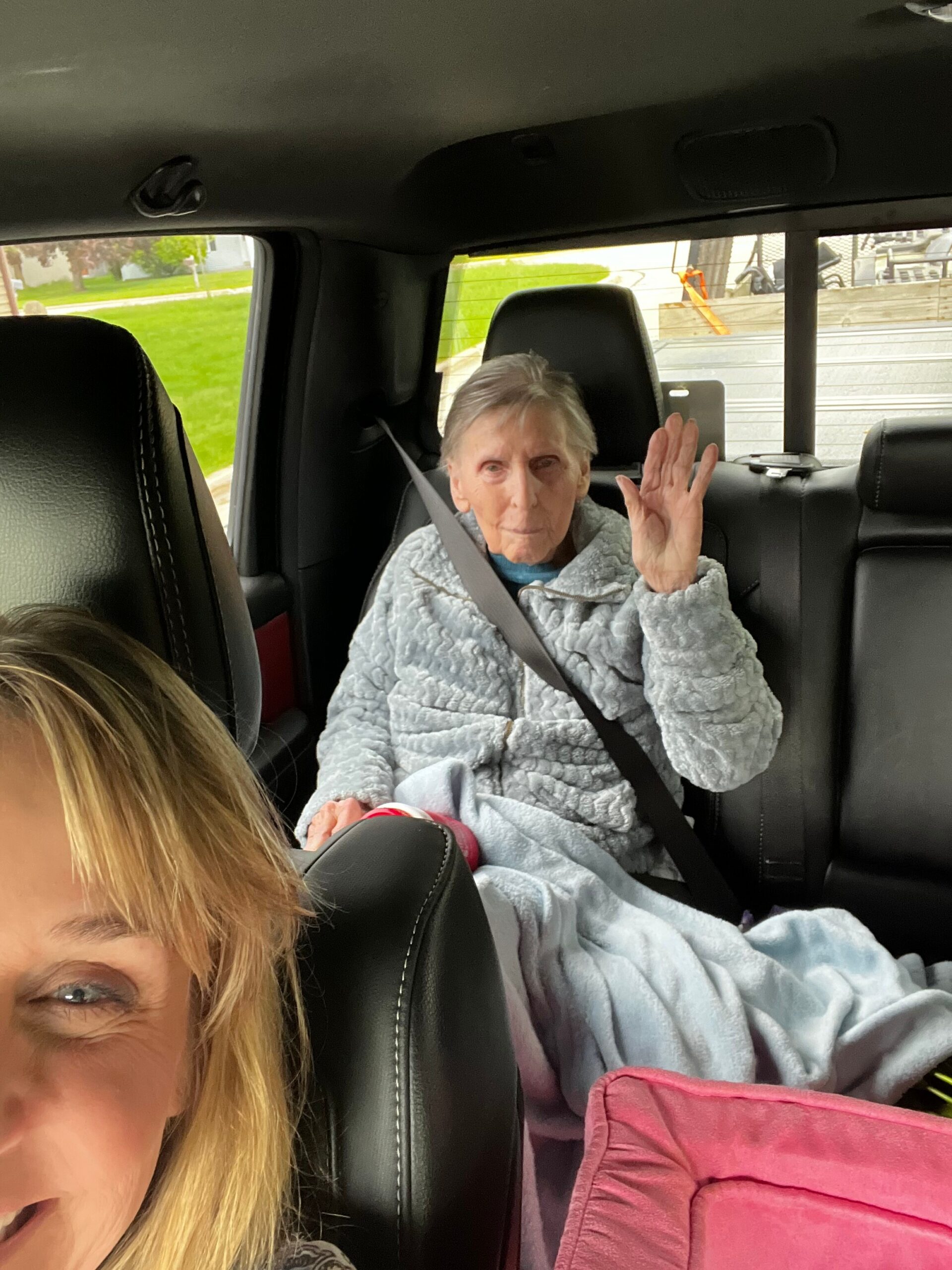 Older woman in a gray robe sits in the back seat of a car, waving at the camera with a seatbelt across her chest, while a foreground passenger is partially visible.