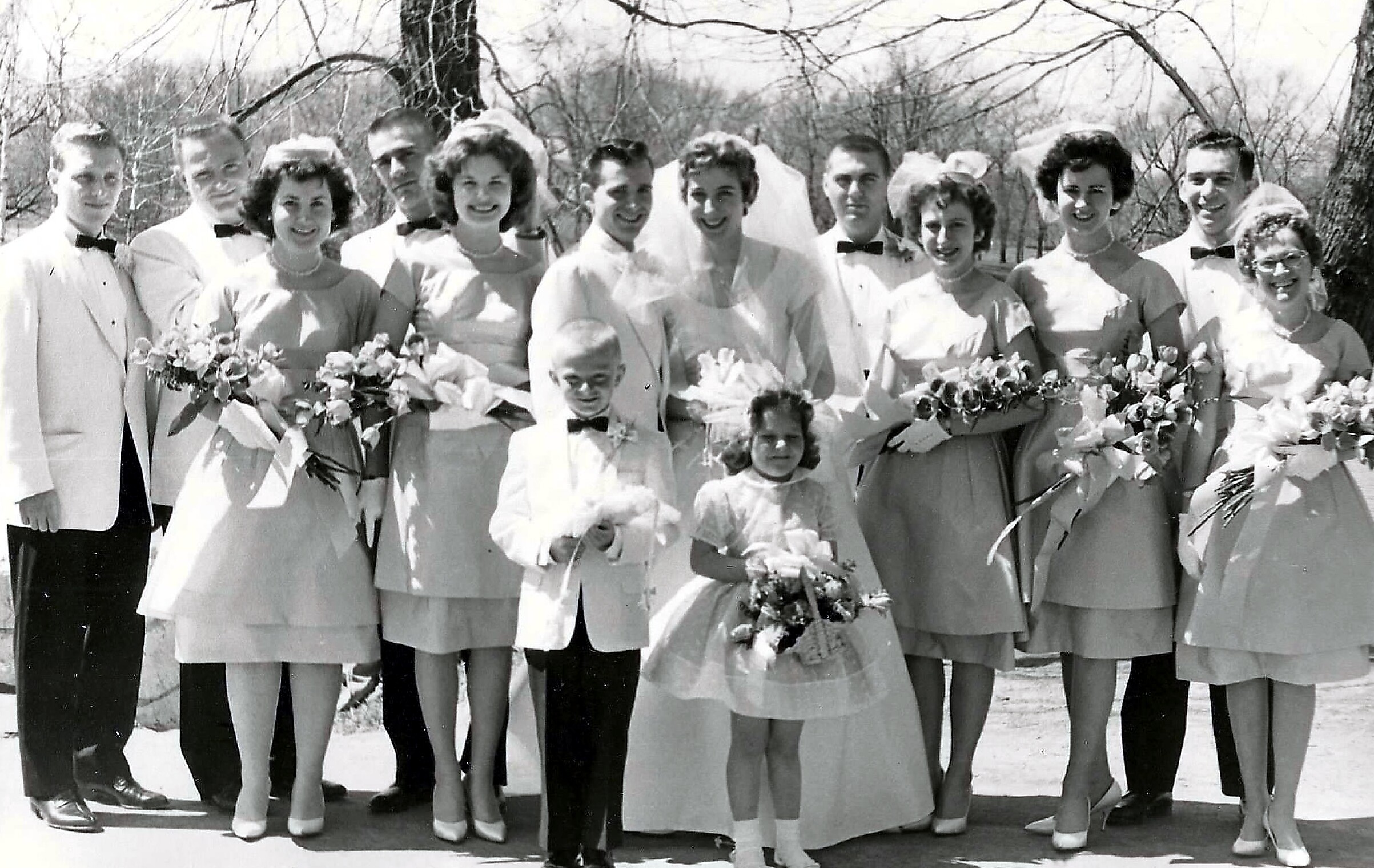 Outdoor black-and-white wedding party photo: bridesmaids in pale dresses with bouquets, groomsmen in white jackets, two children in front.