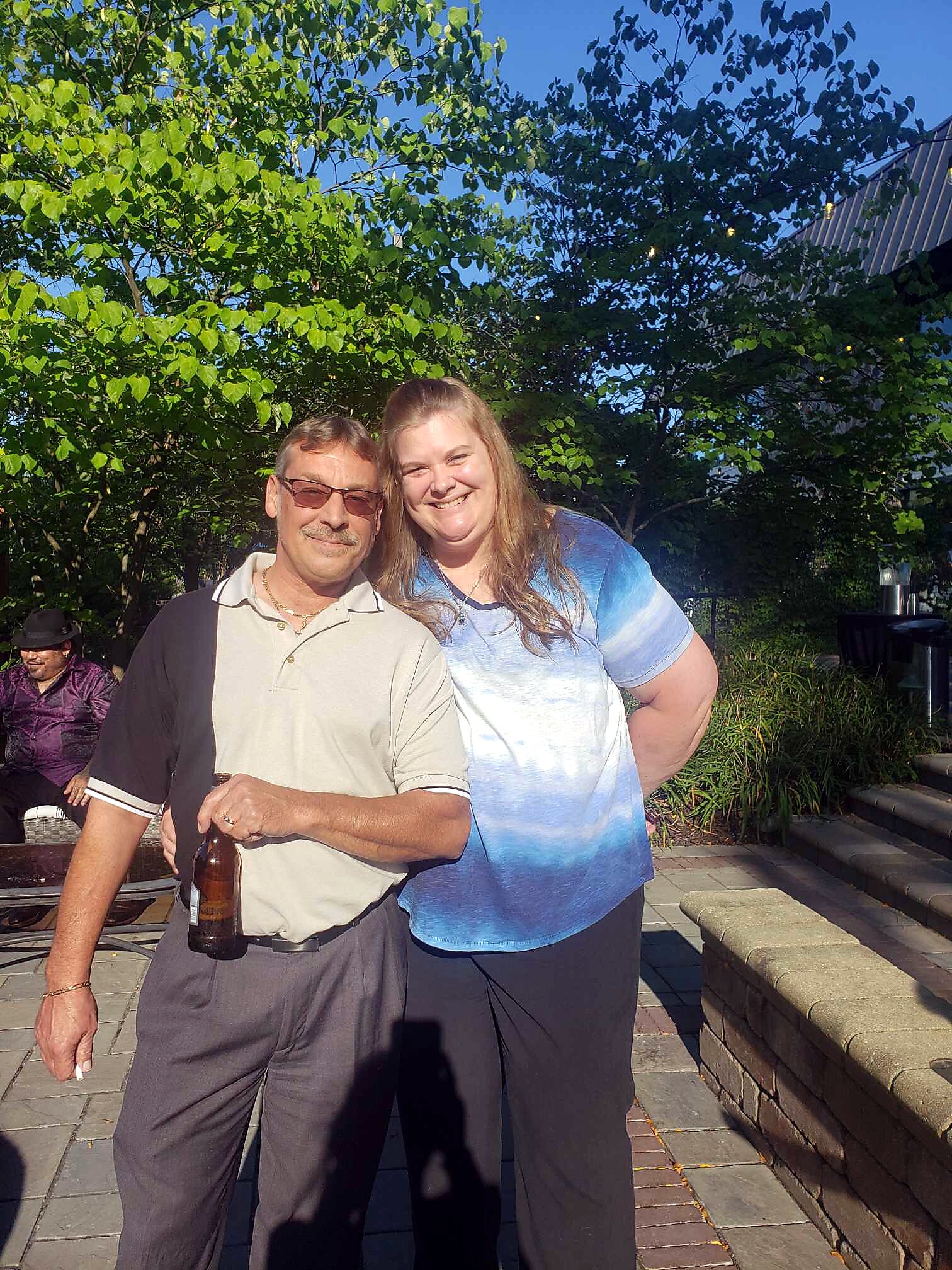 Smiling couple posing outdoors near green trees on a sunny day; man holds a beer bottle, woman leans in.