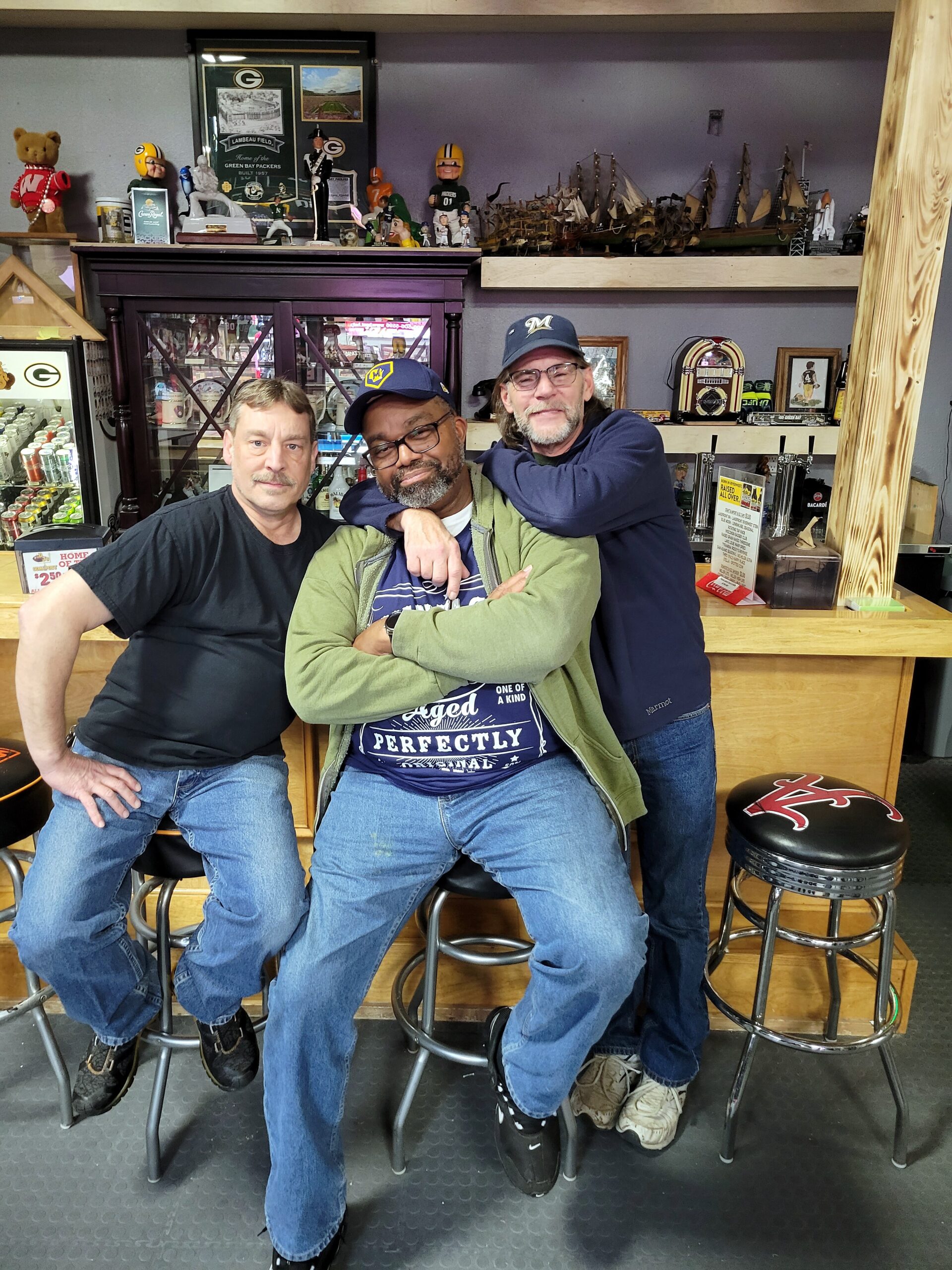 Three men pose with arms around each other in a memorabilia shop filled with model ships and collectibles behind them.