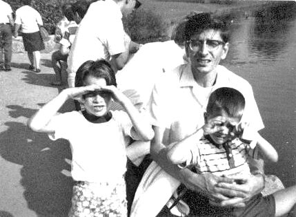 Man crouching by a river with two children; the girl shades her eyes from the sun while the younger boy sits on his lap, with others in the background.
