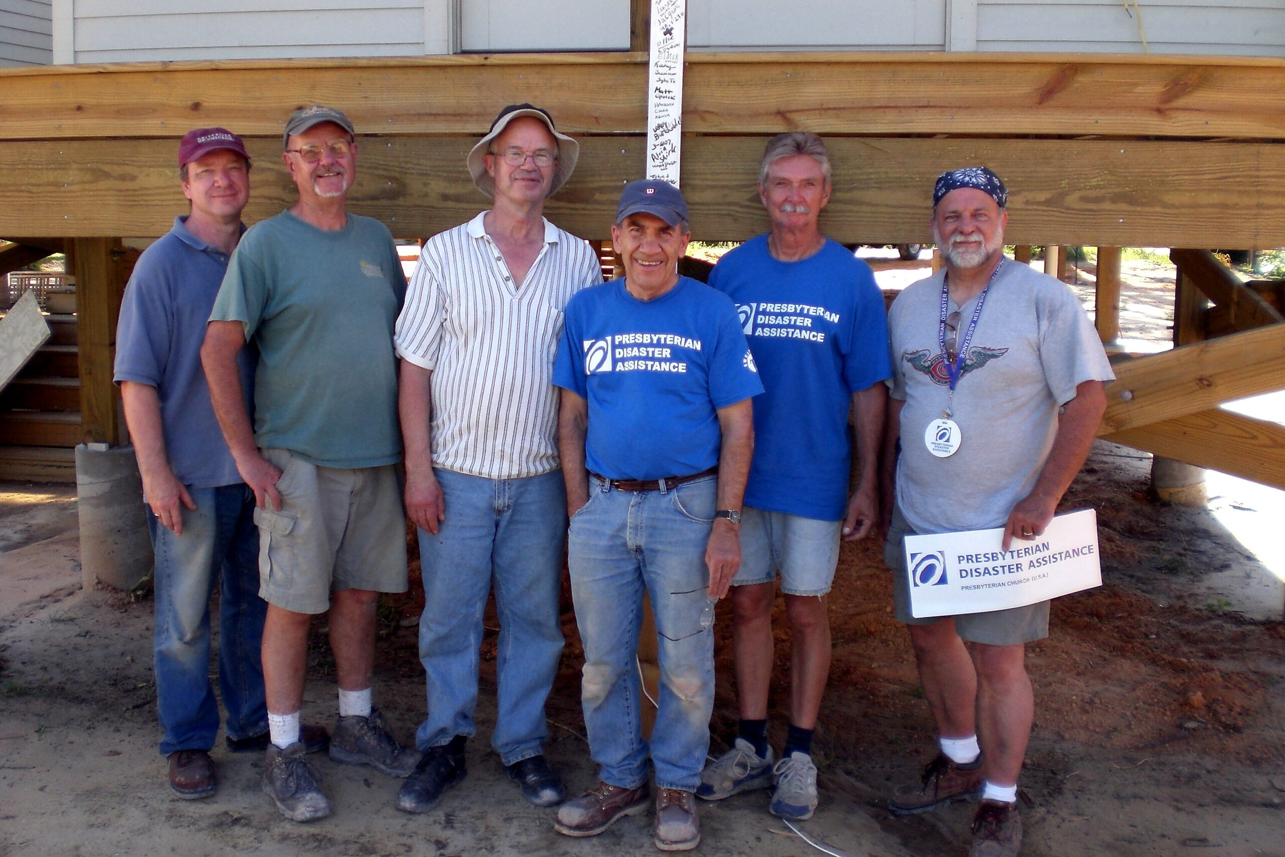 Six volunteers stand together in front of a wooden structure, some wearing blue Presbyterian Disaster Assistance shirts, on a construction site.