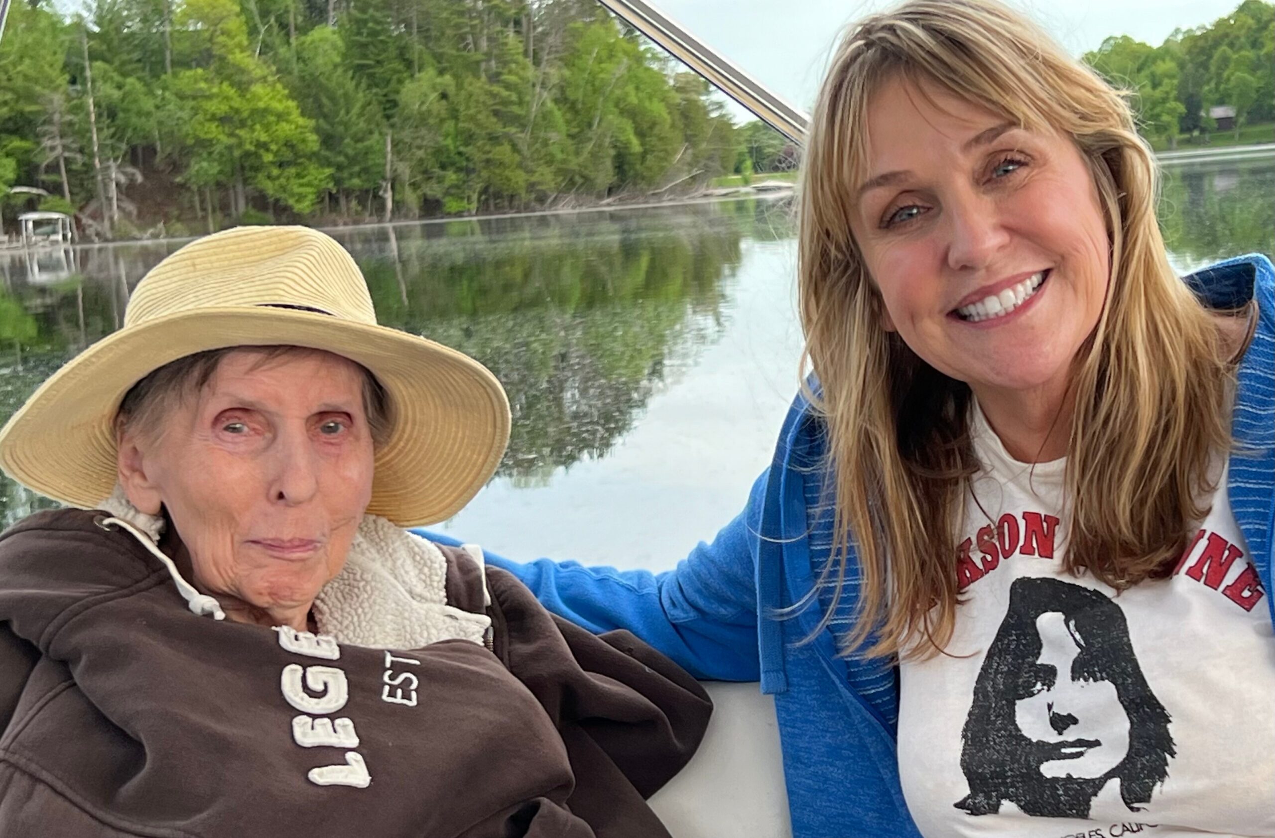 Two women sit together on a boat by a calm lake, smiling at the camera with a forested shoreline behind them.