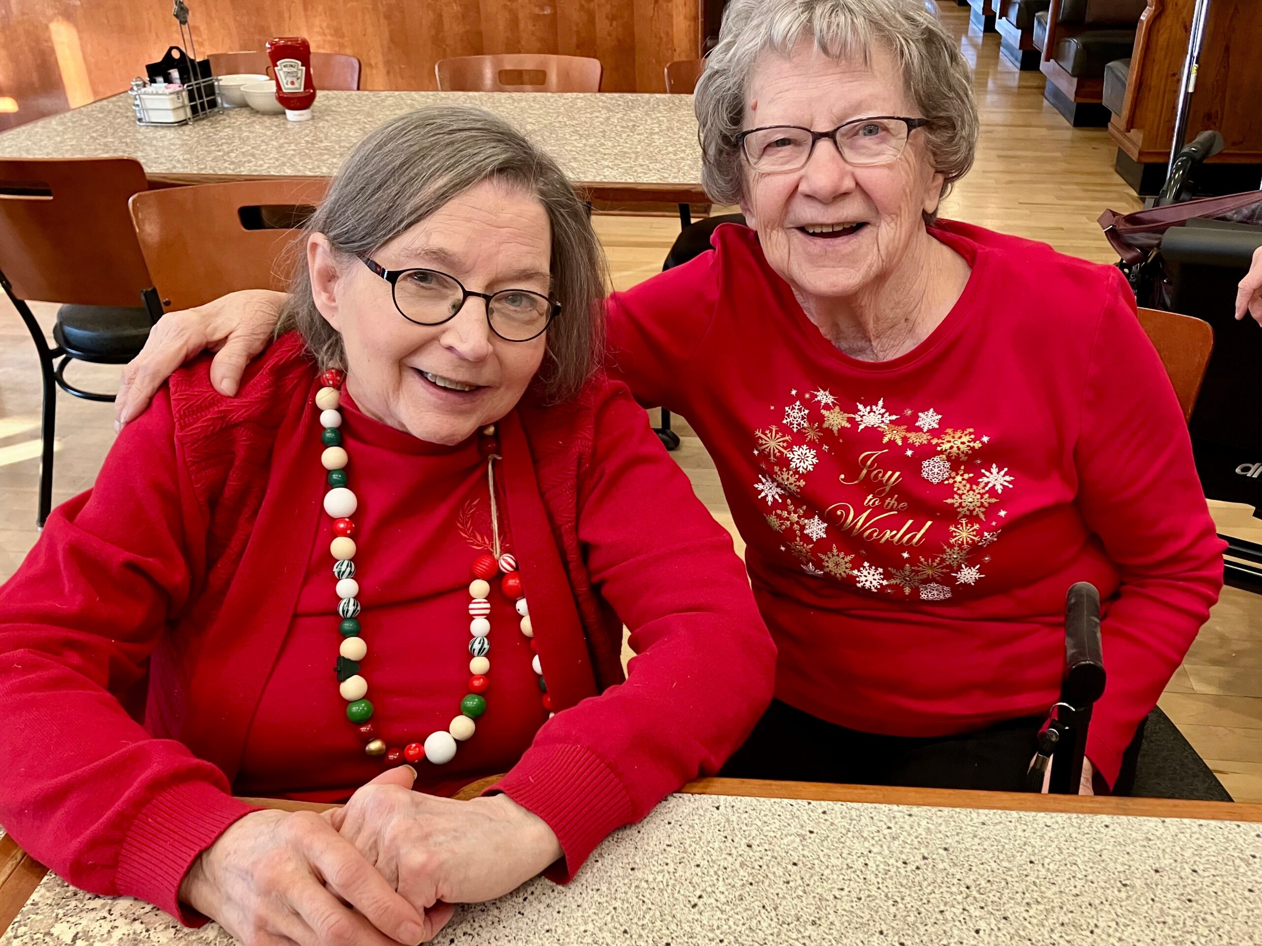 Two elderly women in red festive outfits smile at the camera while seated at a diner table; one wears a bead necklace and the other a snowflake design shirt, ketchup on the table behind them.