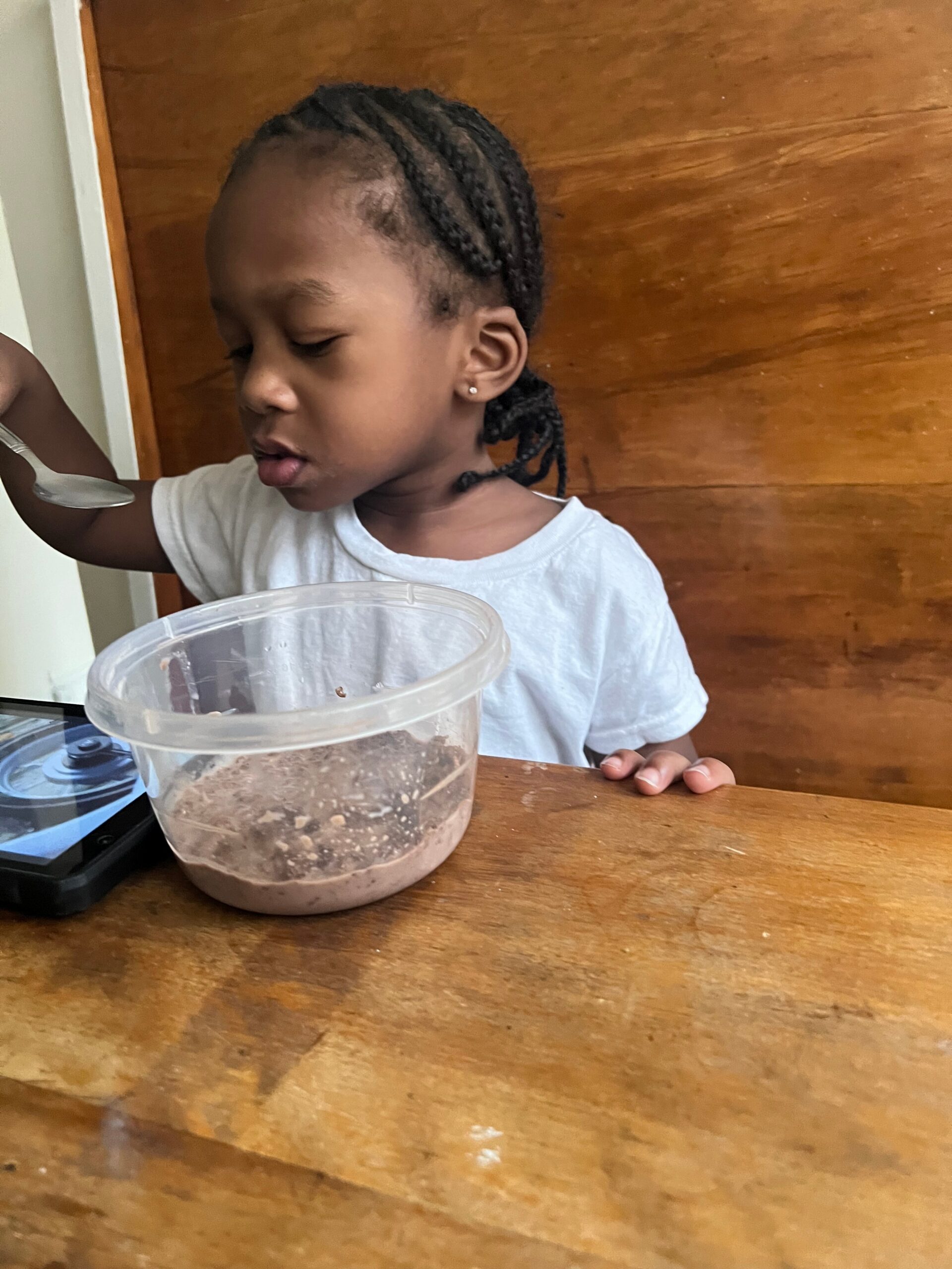 Young child with braided hair sits at a wooden table, looking at a clear container of food with a spoon nearby.