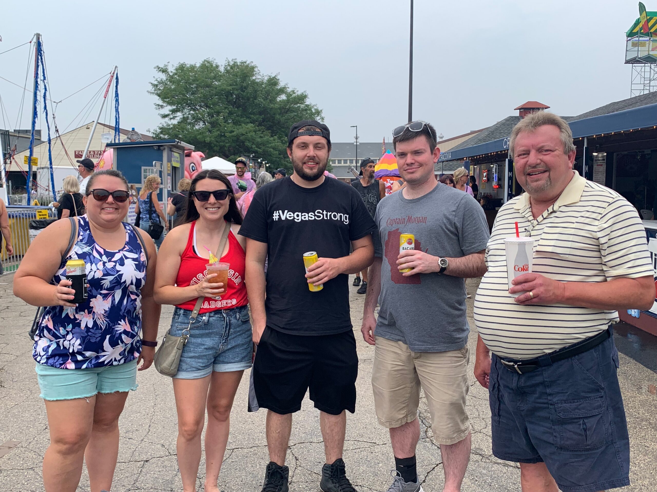 Five adults posing for a group photo at an outdoor fair, each holding drinks and smiling at the camera.],