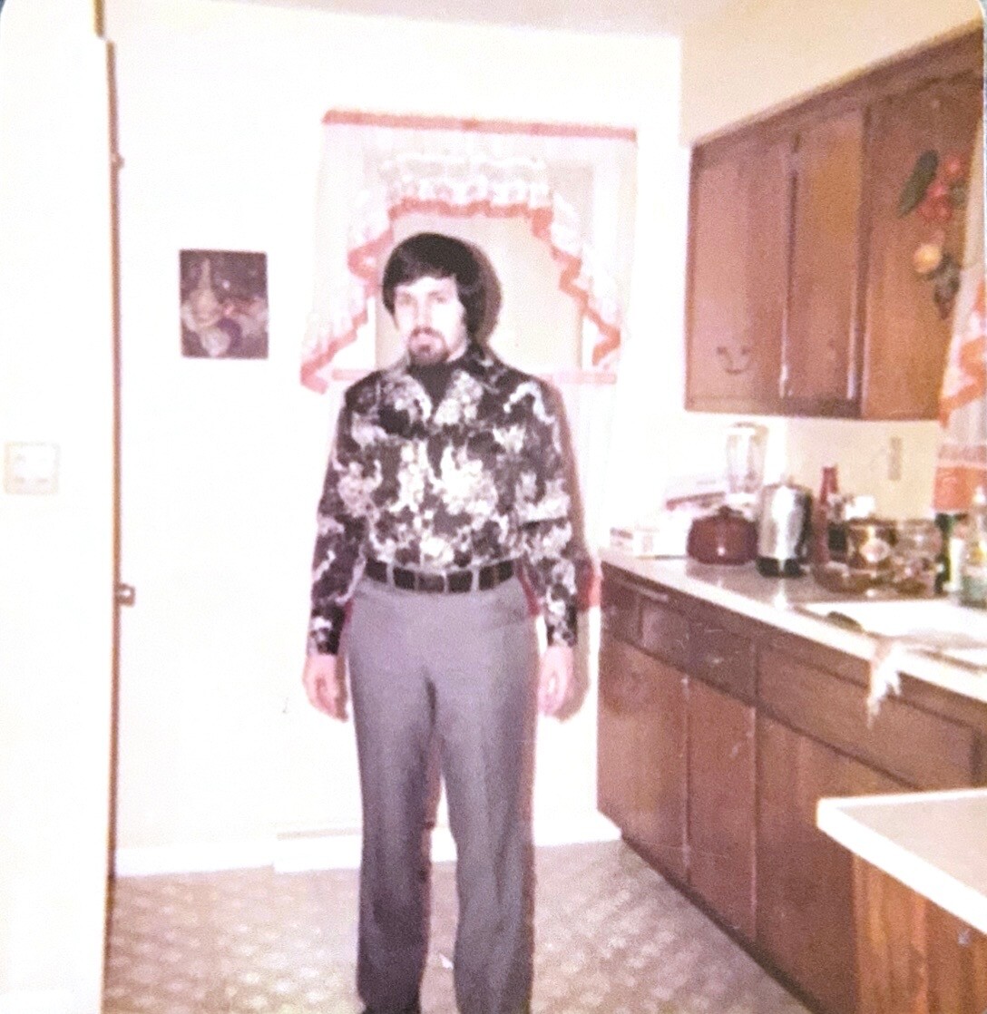 Man in a black-and-white floral shirt and gray pants stands in a retro-style kitchen with wooden cabinets behind him.