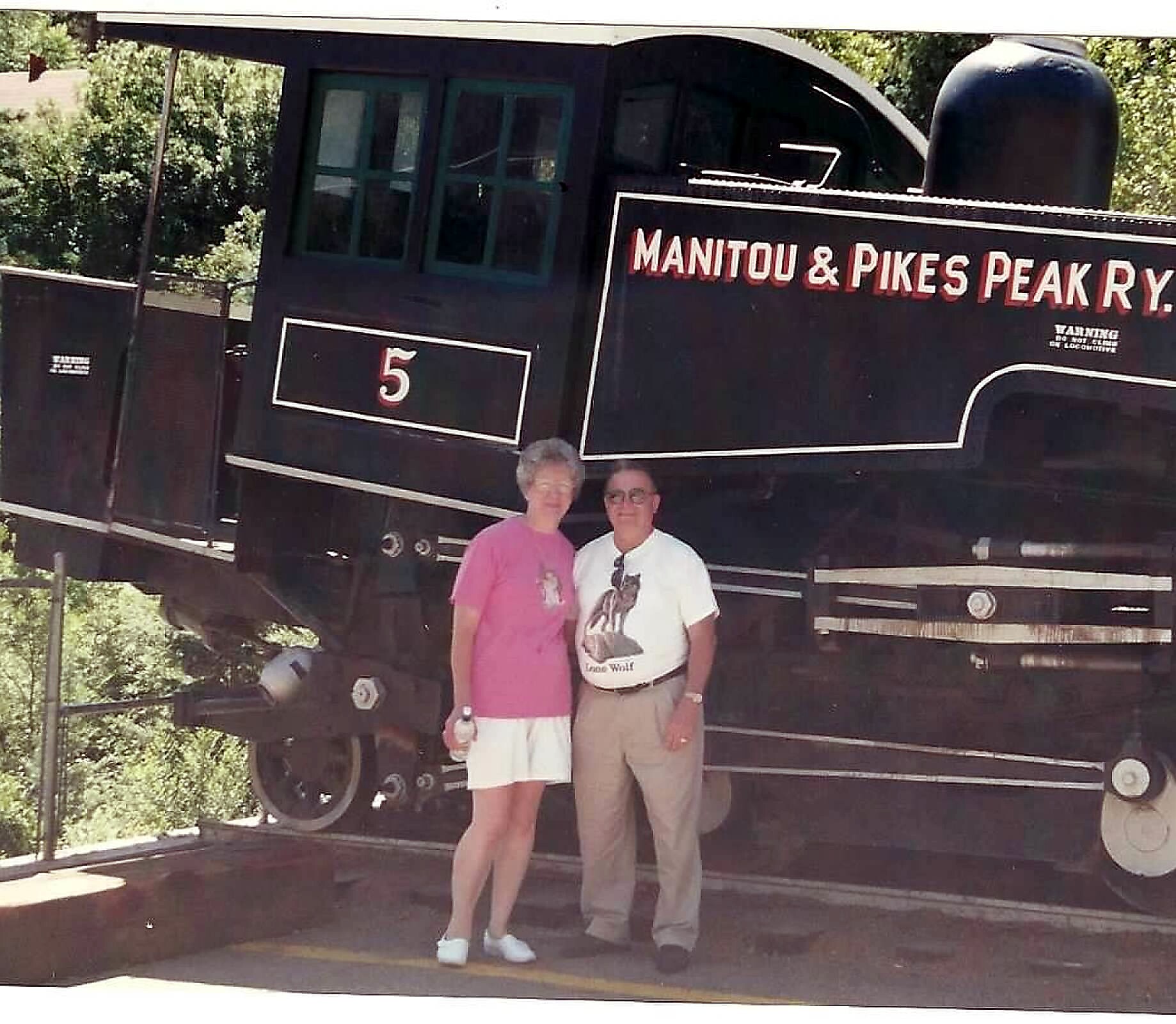 Two people pose in front of a large vintage steam locomotive labeled Manitou & Pike's Peak R.R.