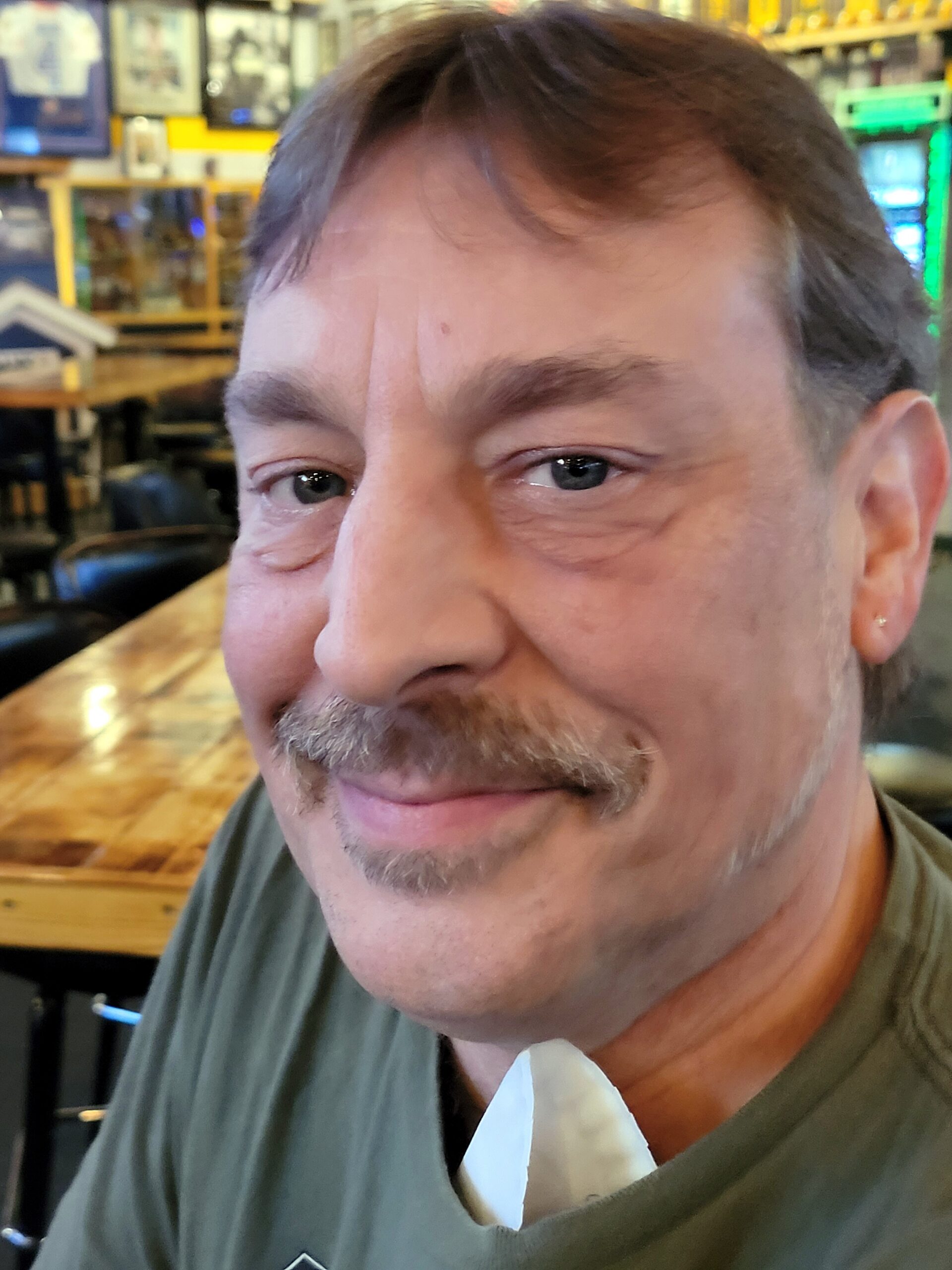 Close-up of a smiling man with a brown mustache in a casual café setting, blurred background.