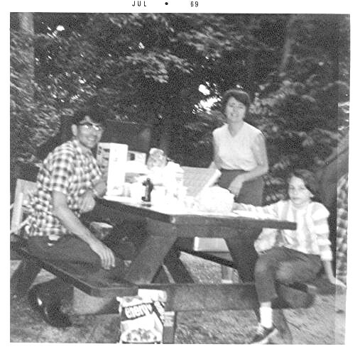 Family of four at an outdoor picnic table in a backyard, with food on the table, posing for a photo.