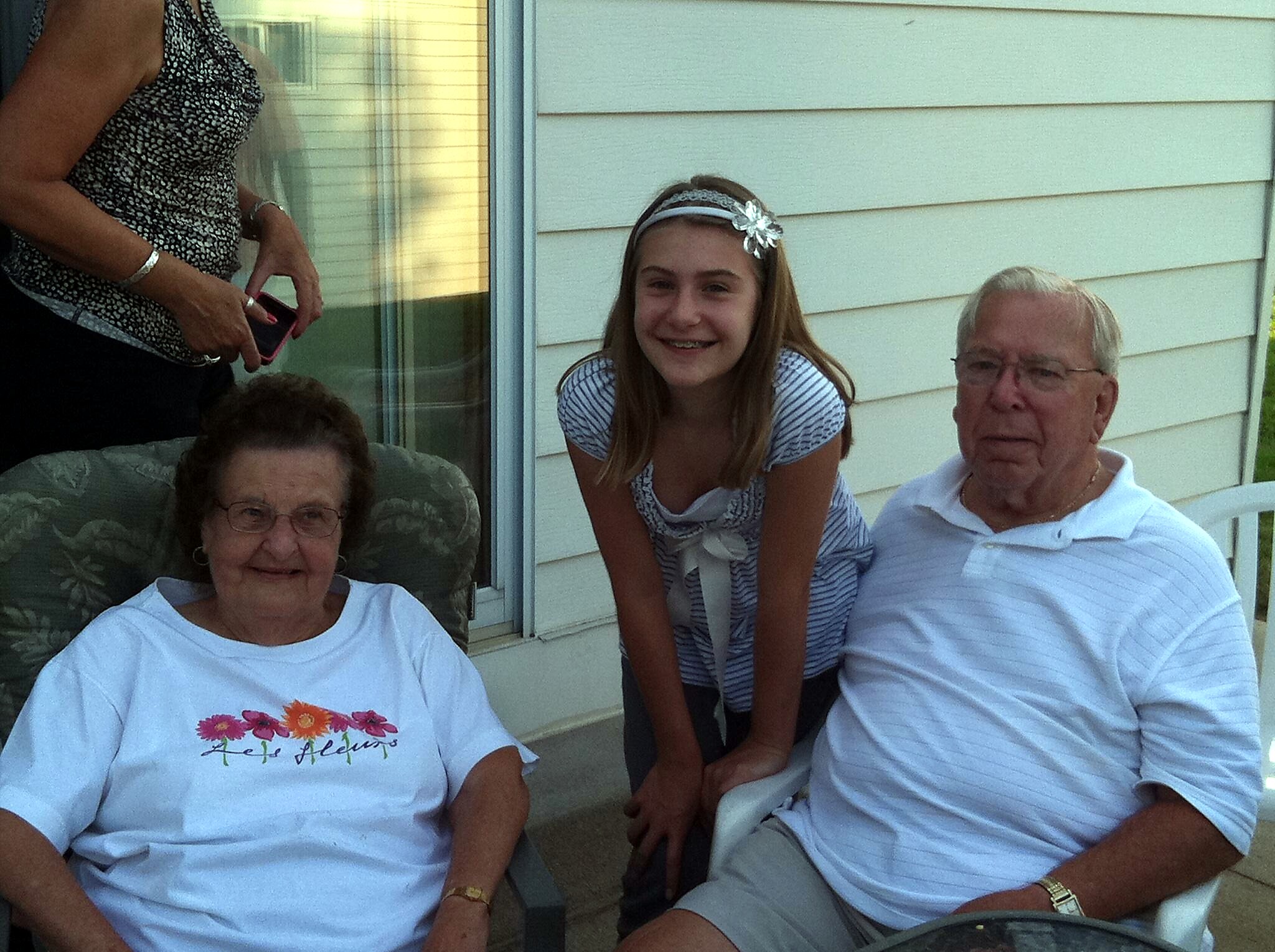 Three people posing on a sunny porch: an older woman seated left, a smiling teenage girl leaning forward in the middle, and an older man seated right.