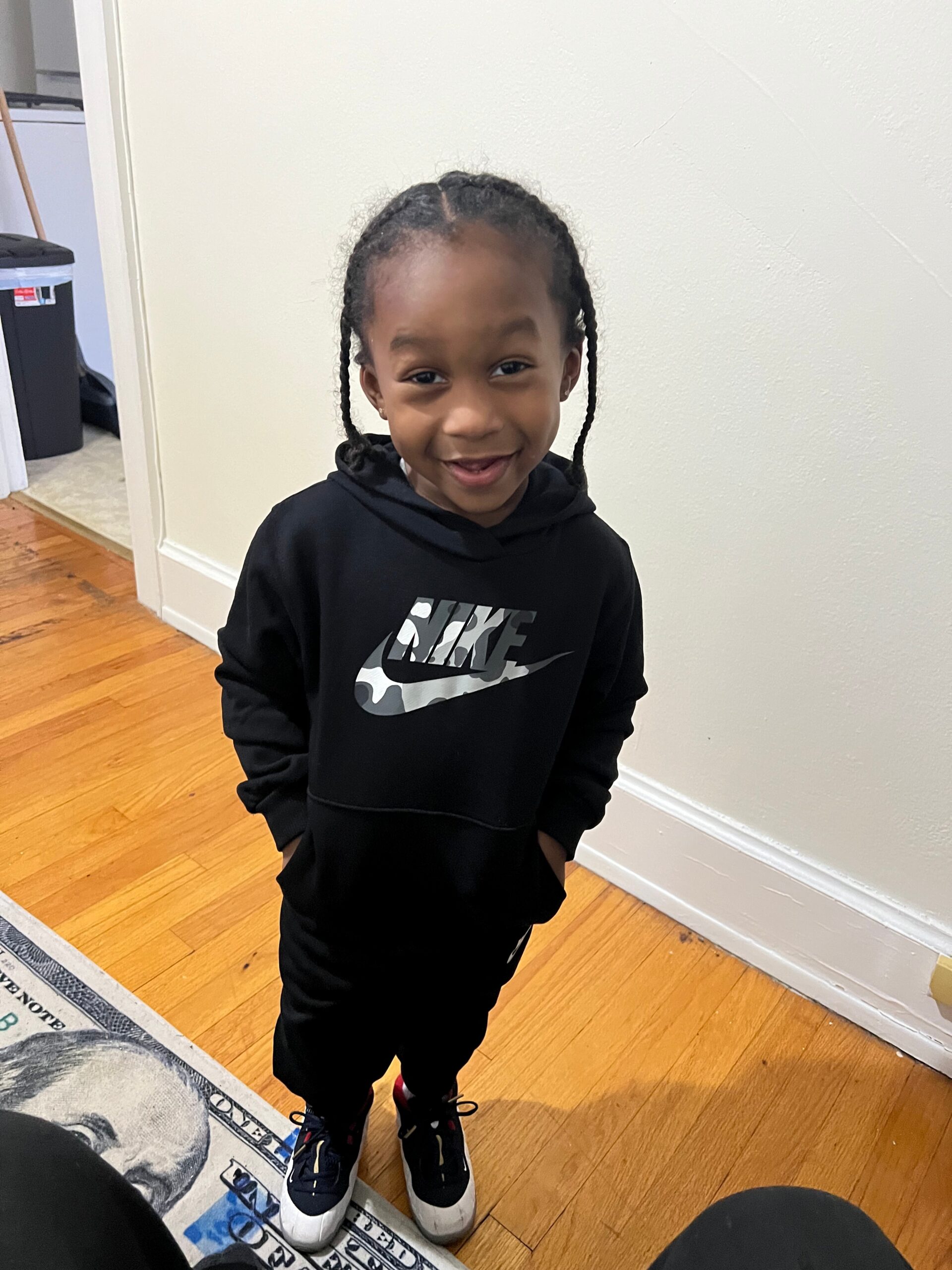A young girl with braided hair standing indoors, wearing a black Nike hoodie and sneakers, smiling at the camera.