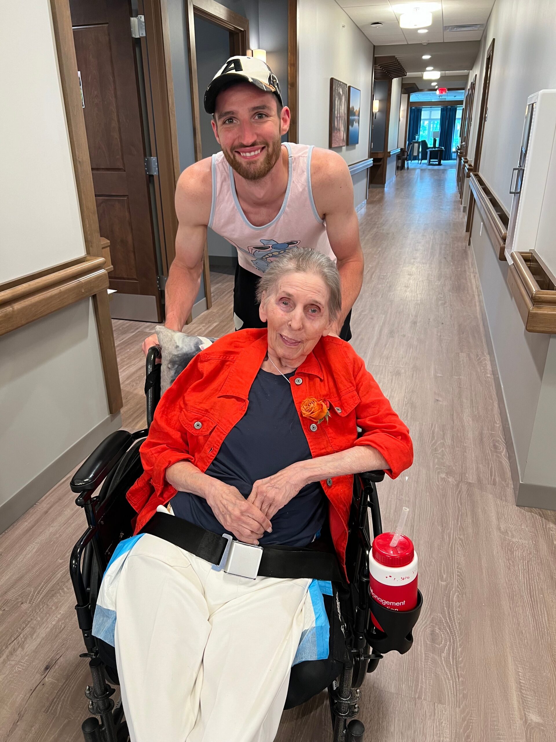 Smiling man pushes an elderly woman in a wheelchair down a hospital-like hallway.