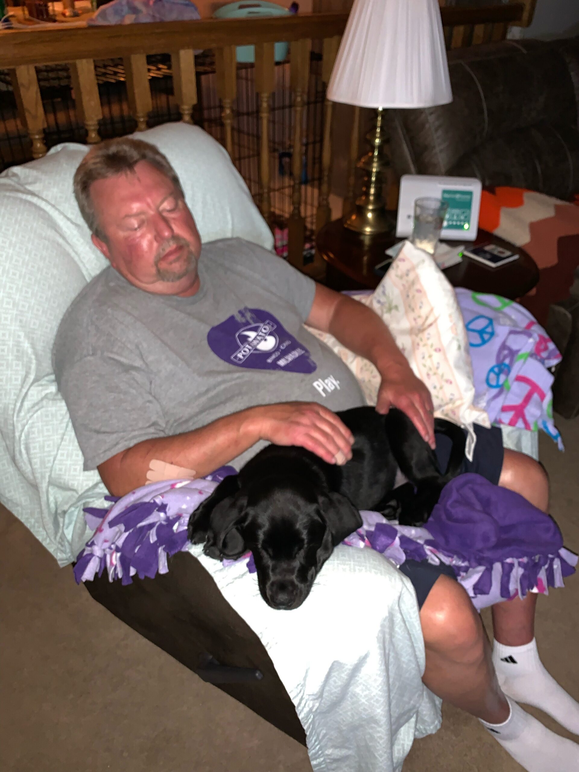 Older man sleeping on a recliner with a black dog resting on his lap, surrounded by blankets and a table lamp nearby in a cozy living room.