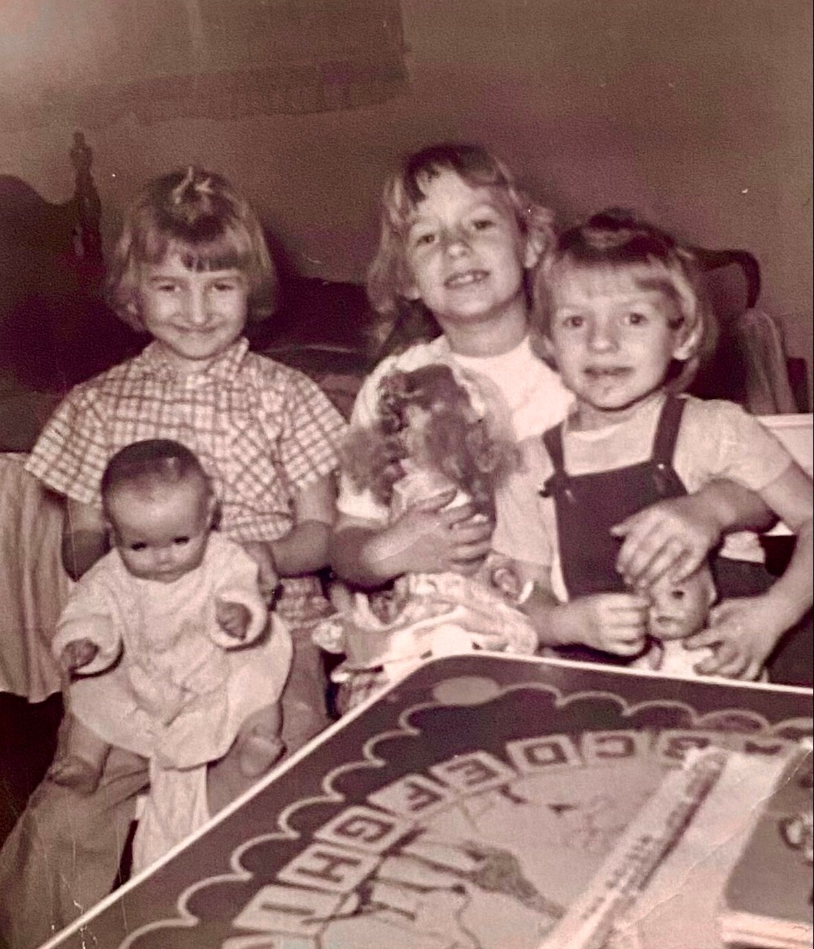 Four young children pose on a couch in a vintage photo: a baby in front left, two older girls in the middle, and a girl on the right with a doll.”,