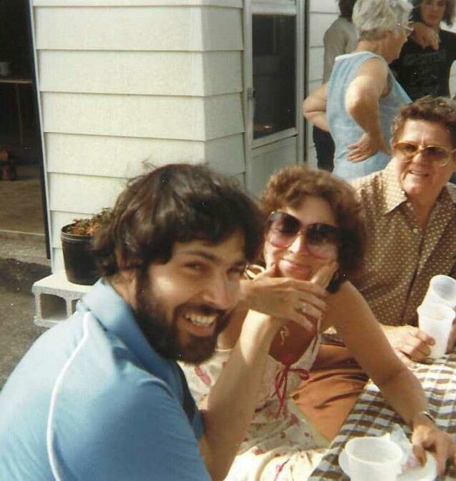Man with dark hair and beard smiling at the camera, seated with a woman in sunglasses at a checkered table during an outdoor gathering.