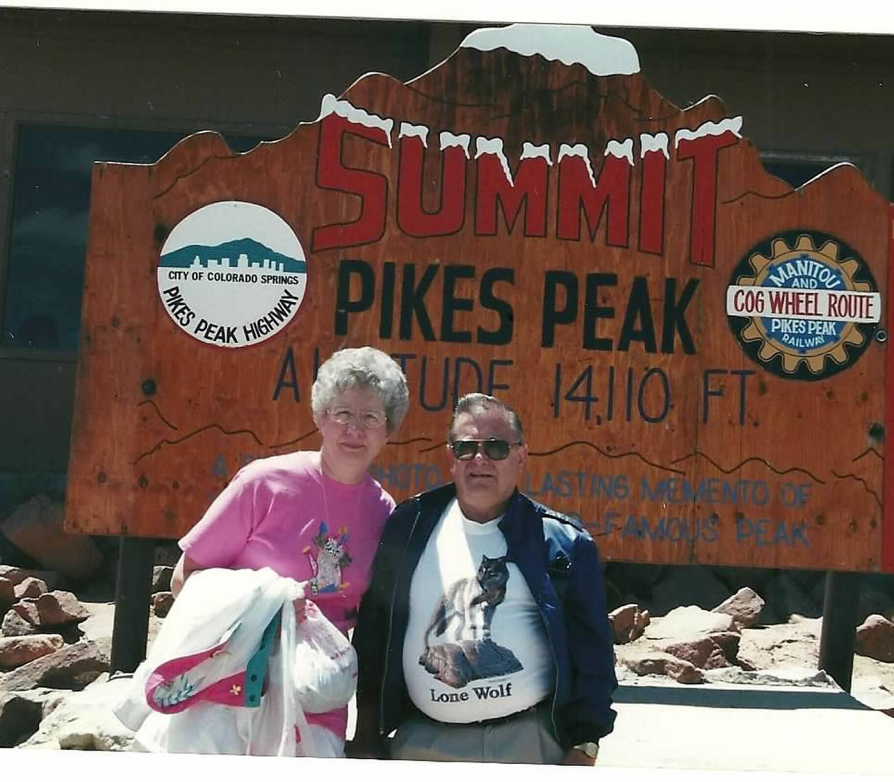 Two people stand in front of a large orange Summit sign at Pikes Peak, Colorado Springs, with logos on the sign behind them.