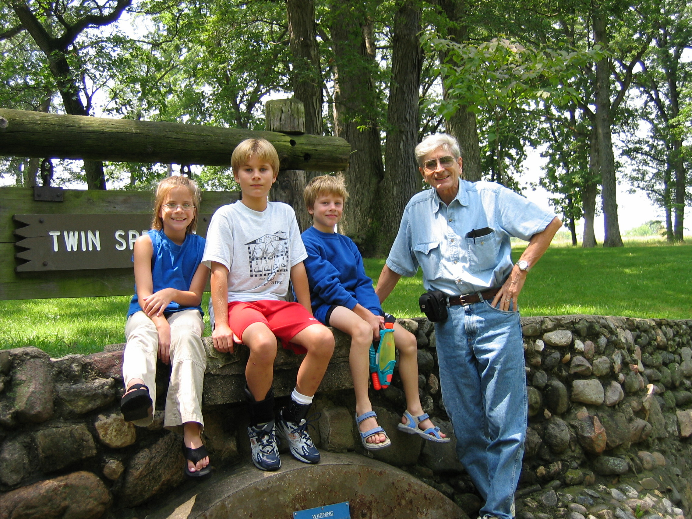 Group of four children and an older man posing on a stone wall in a park, with a wooden sign in the background.