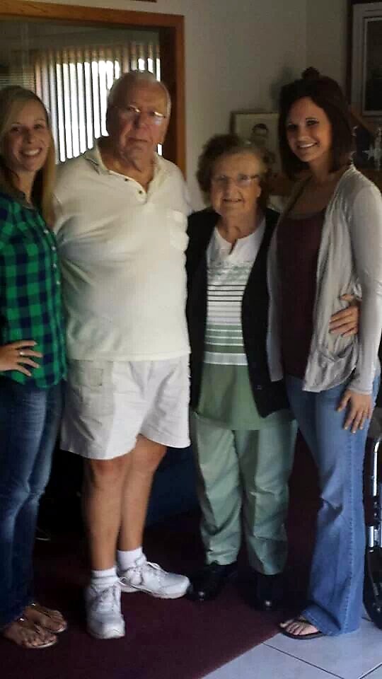 Four people pose for a group photo indoors: two younger women on ends and an elderly couple in the middle.