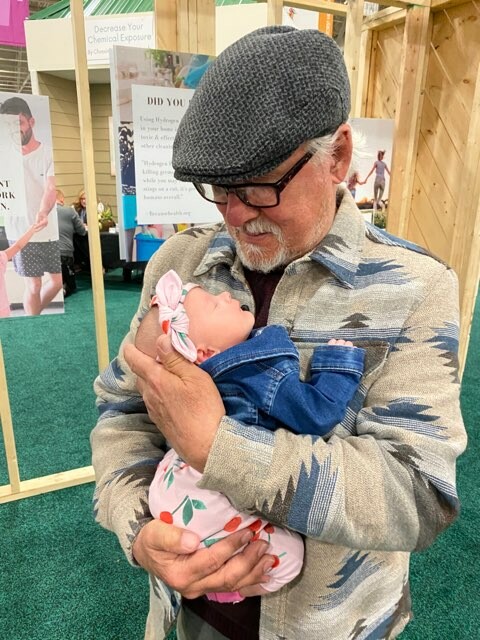 Older man with a gray flat cap and glasses cradles a newborn girl in a pink floral outfit, smiling down at her.